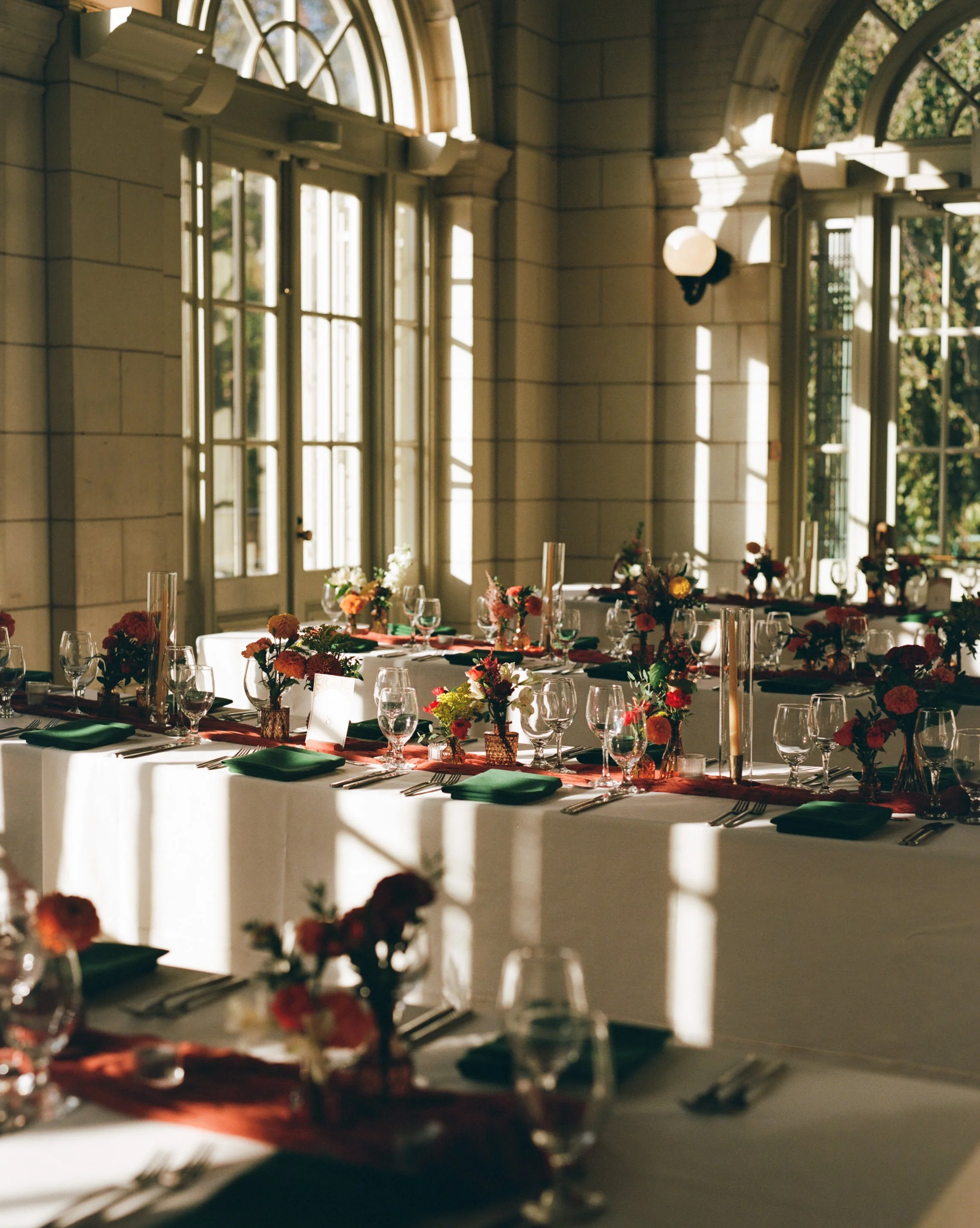 Elegant indoor setting with a long table decorated with flowers, candles, and tableware, illuminated by sunlight through large arched windows in a historic building.