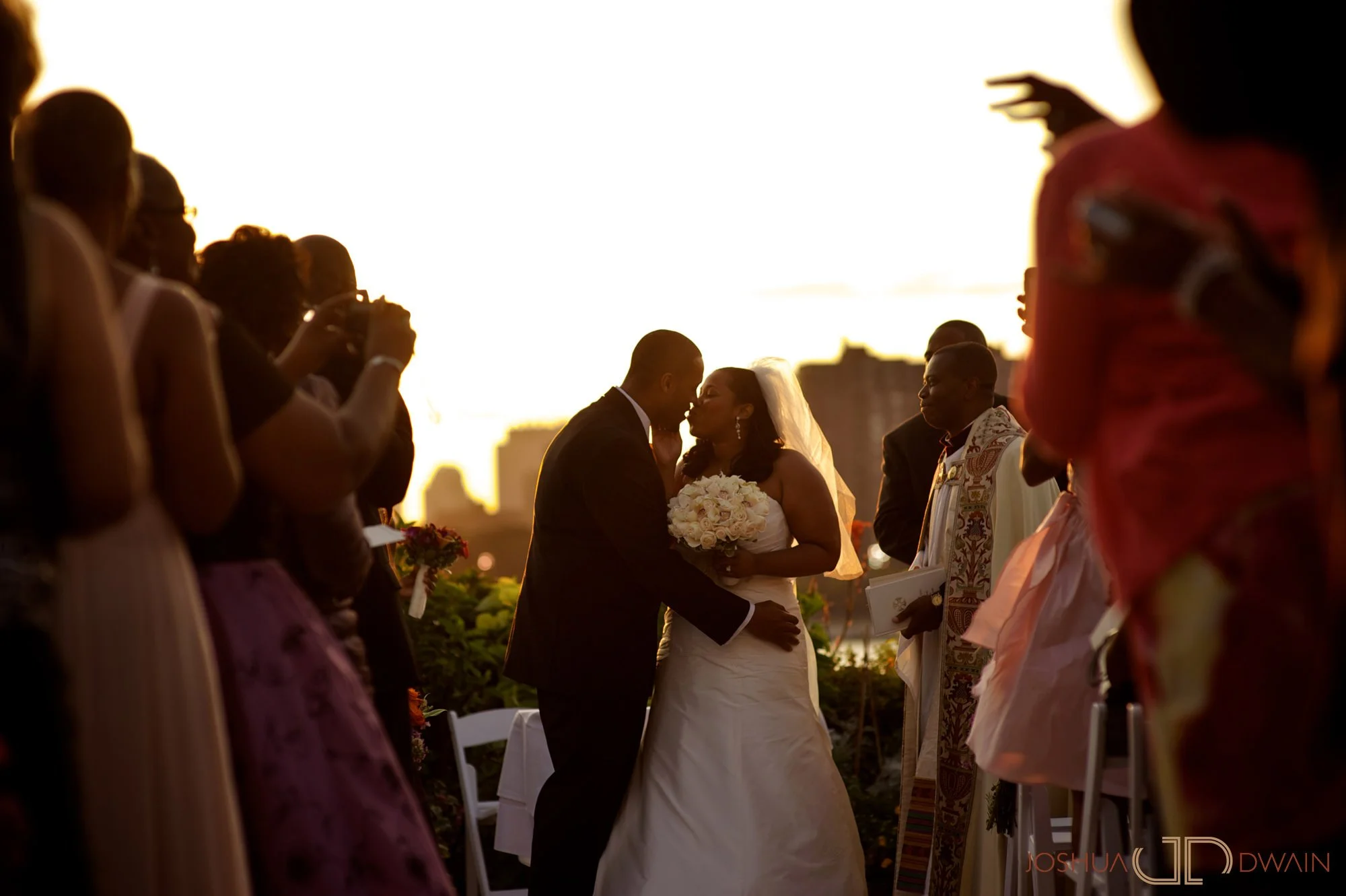 A bride and groom share a kiss during their wedding ceremony at sunset, surrounded by guests in formal attire.