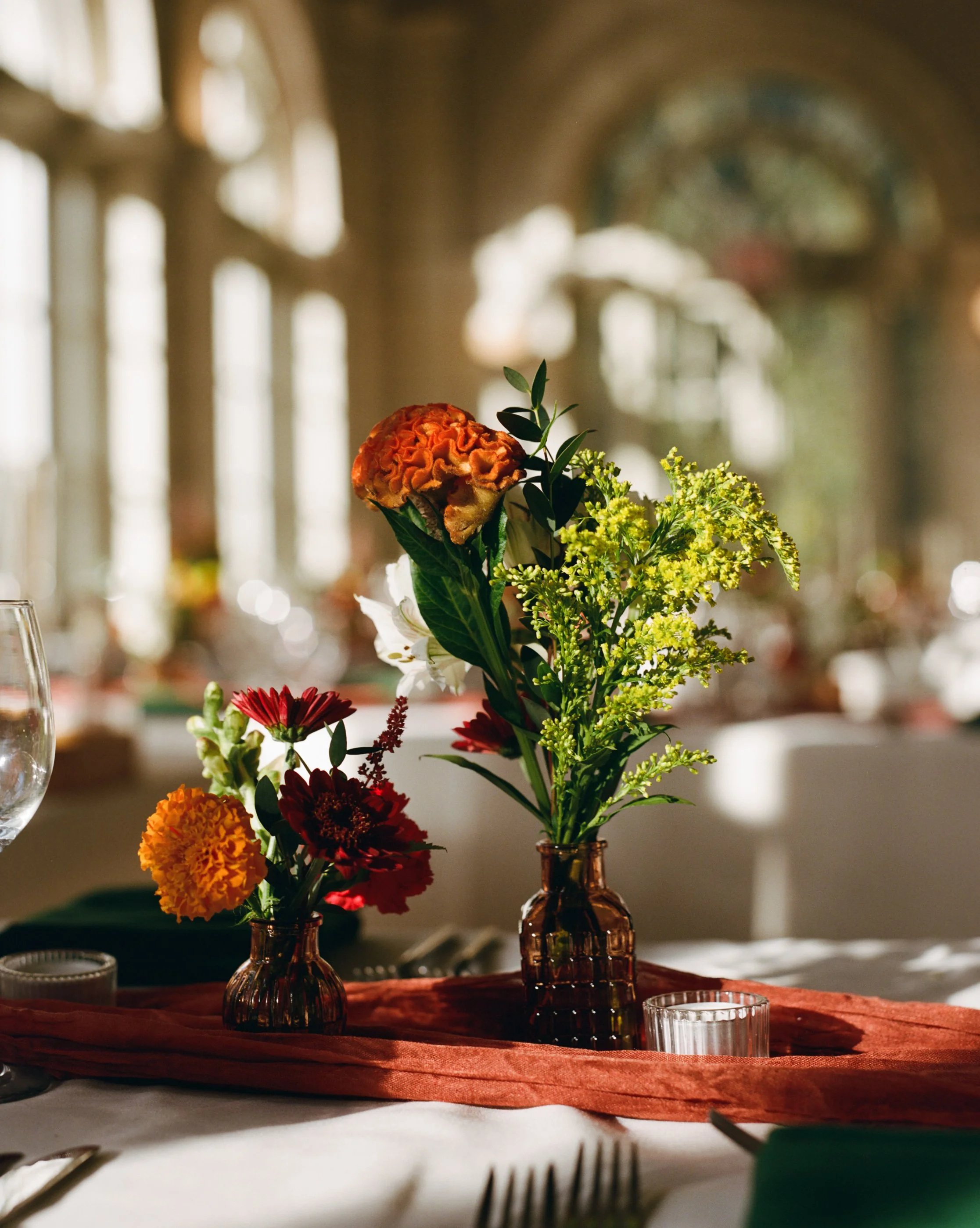 Close-up of a floral centerpiece with orange and red flowers in amber vases on a table with a rust-colored table runner, set for a formal event in a sunlit room with large windows and architectural arches.