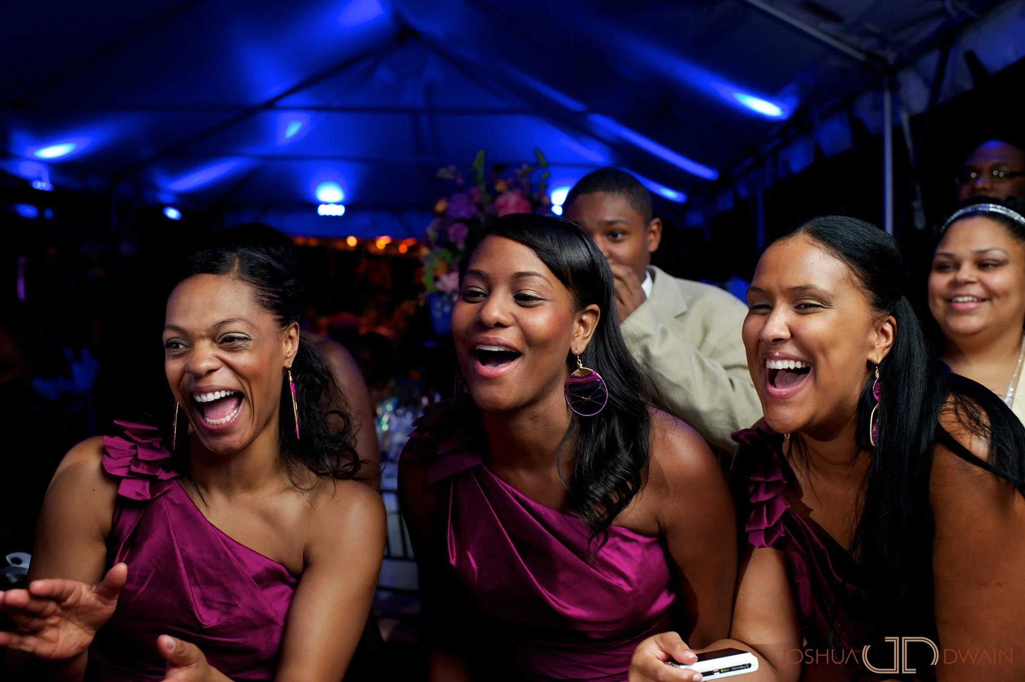 People in vibrant pink dresses laughing and taking photos at an indoor event with blue lighting and floral arrangements in the background.