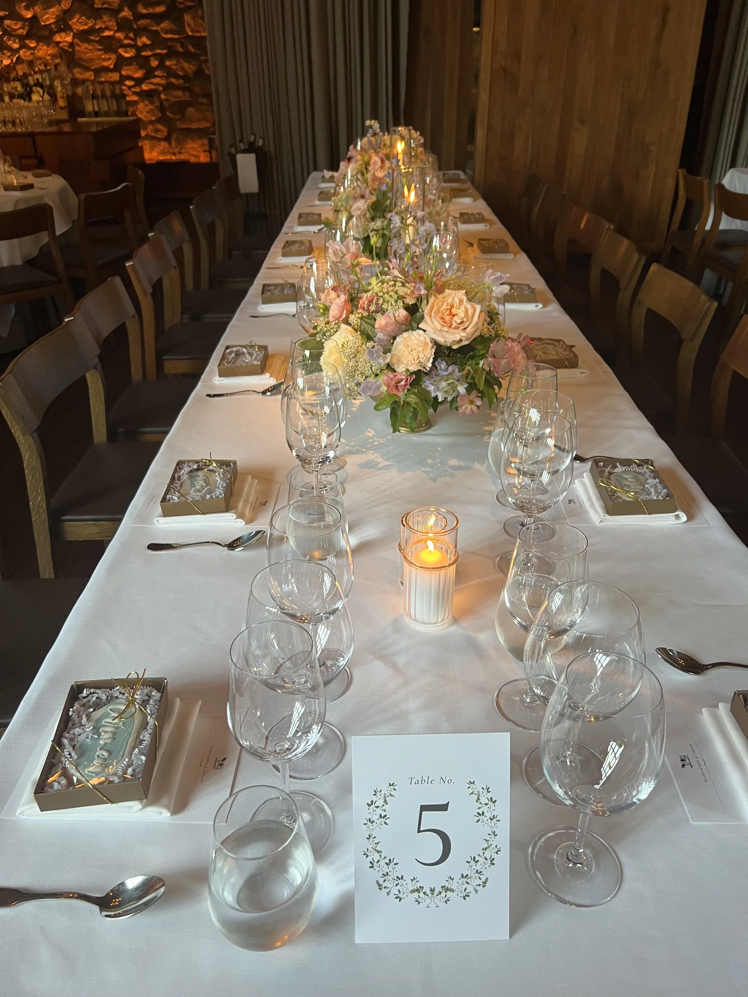 A long dining table decorated with a floral centerpiece, candles, wine glasses, and small gift boxes, set for a wedding reception or formal event. Table number 5 is visible at the front.