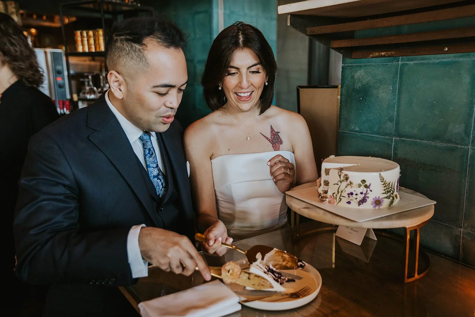 People at a celebration, serving themselves cake at a bakery or cafe, with a decorated cake on a stand nearby.