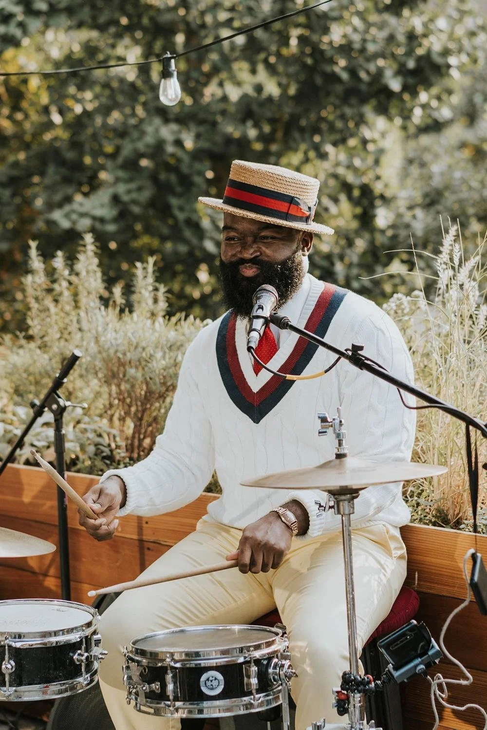 A man with a beard playing drums outdoors, wearing a straw boater hat, a white sweater vest with red and black accents, and beige pants. There are trees and plants in the background, and a string of lights above him.