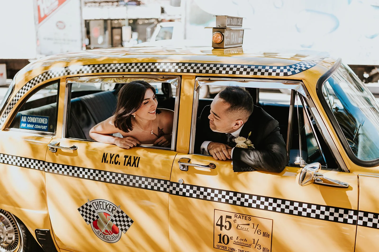 A bride and groom smiling at each other inside a yellow taxi cab titled 'N.Y.C. Taxi'.