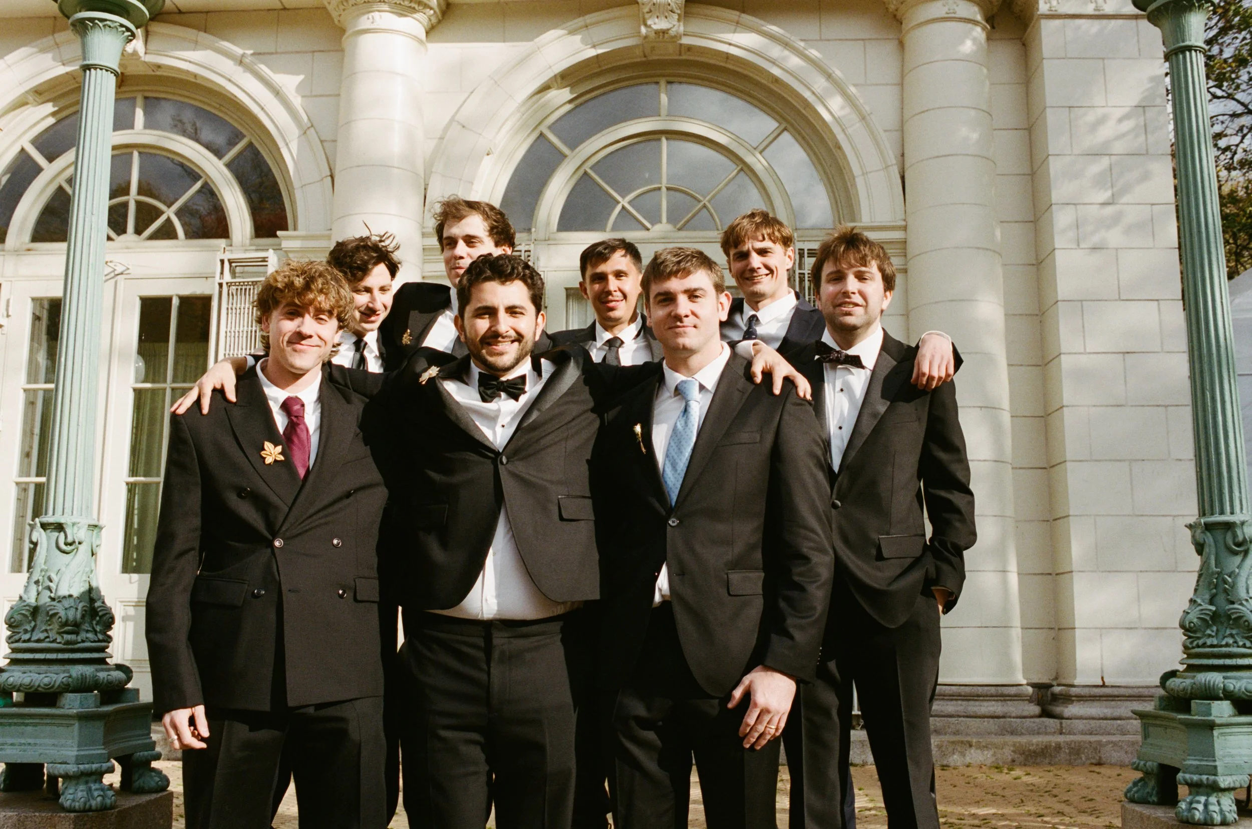 A group of nine young men in tuxedos standing together outside a historic building with large arched windows and columns, smiling and posing for a photo.