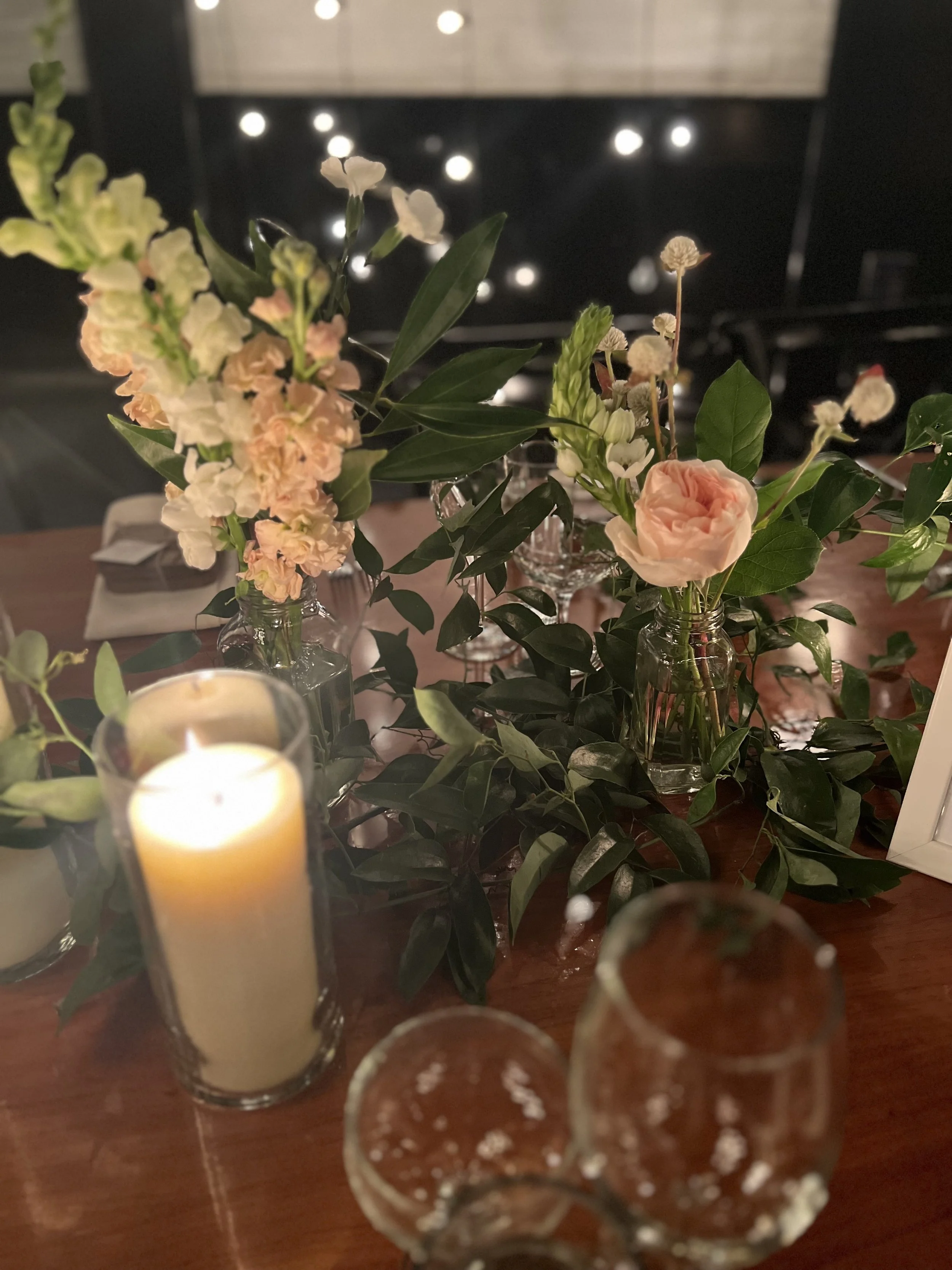 Close-up of floral centerpieces on a table with candles, featuring pink roses, white and pink snapdragons, and greenery, in a dimly lit setting.