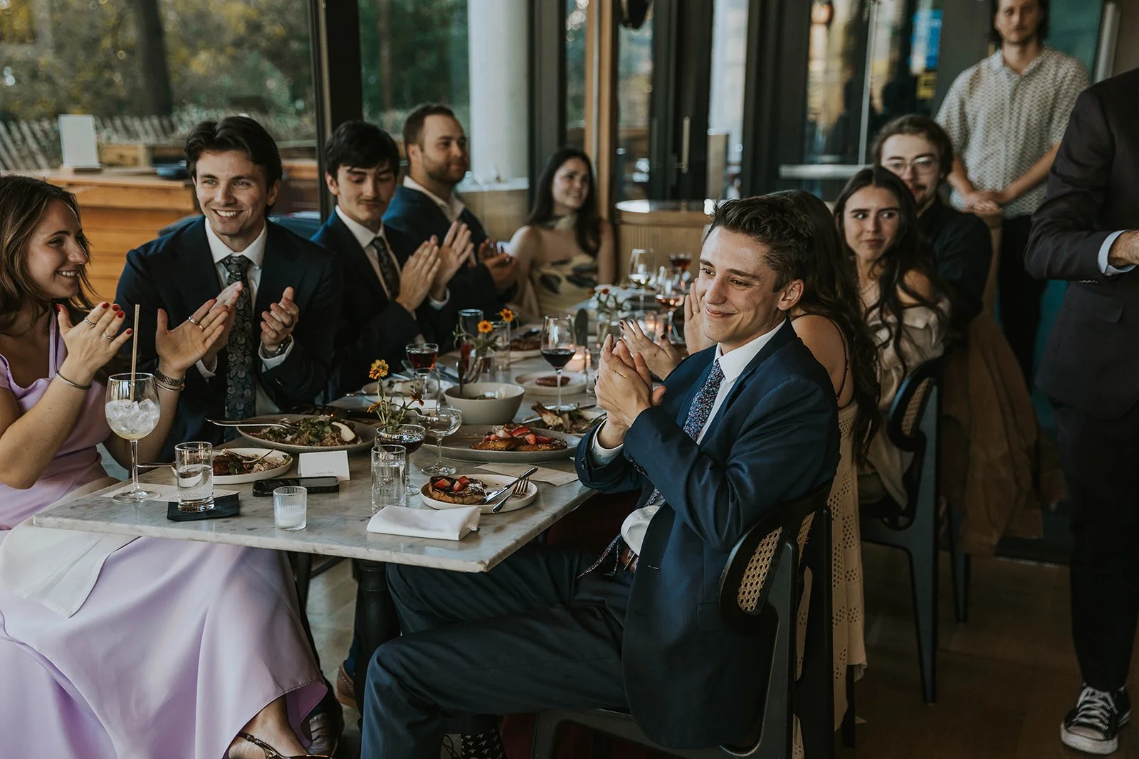 People gathered around a dining table at a celebration, clapping and smiling, with food, drinks, and flowers on the table.