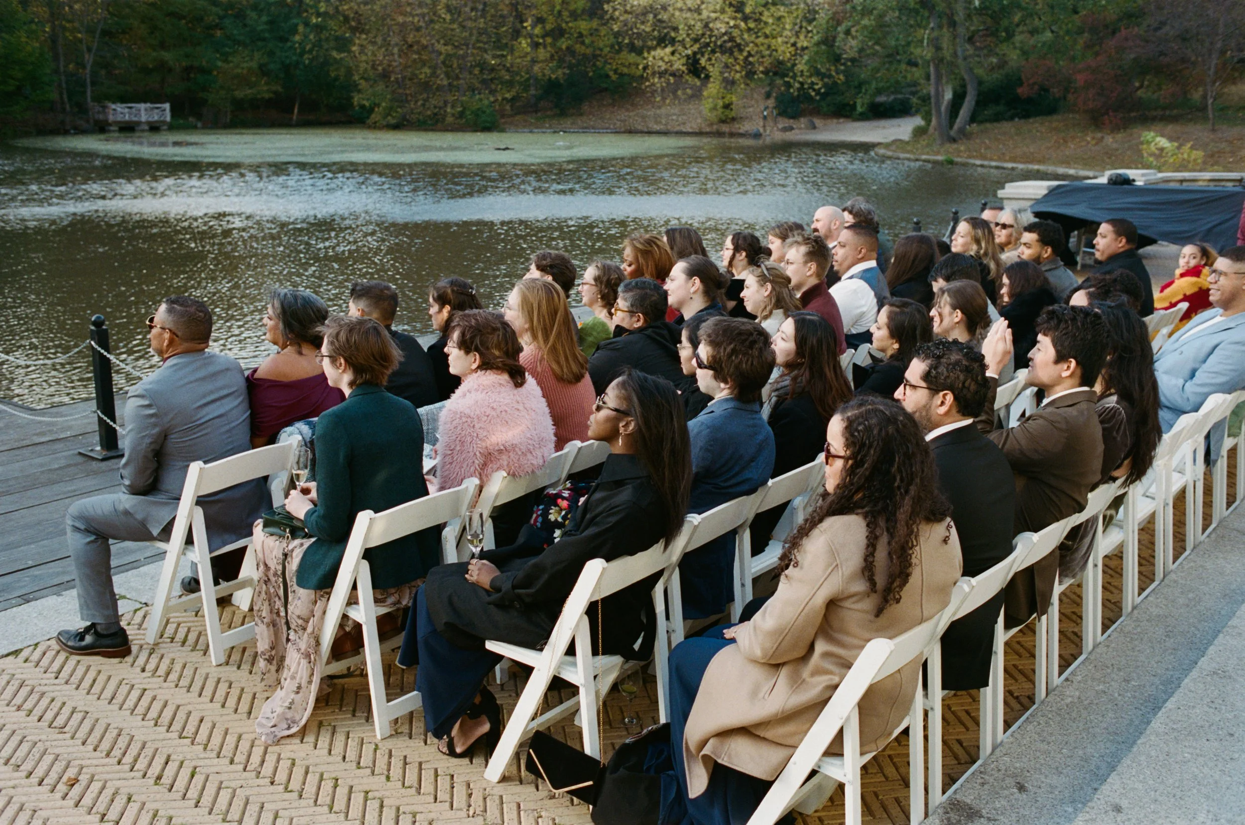People attending an outdoor event beside a lake surrounded by trees during fall, sitting on white chairs on a wooden deck, facing the lake.
