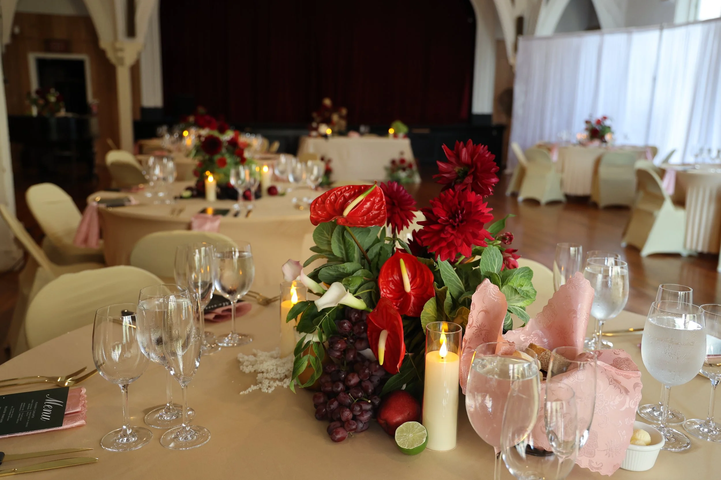 A floral centerpiece with red and white flowers, grapes, candles, and pink napkins on a banquet table set for a formal event in a decorated hall.