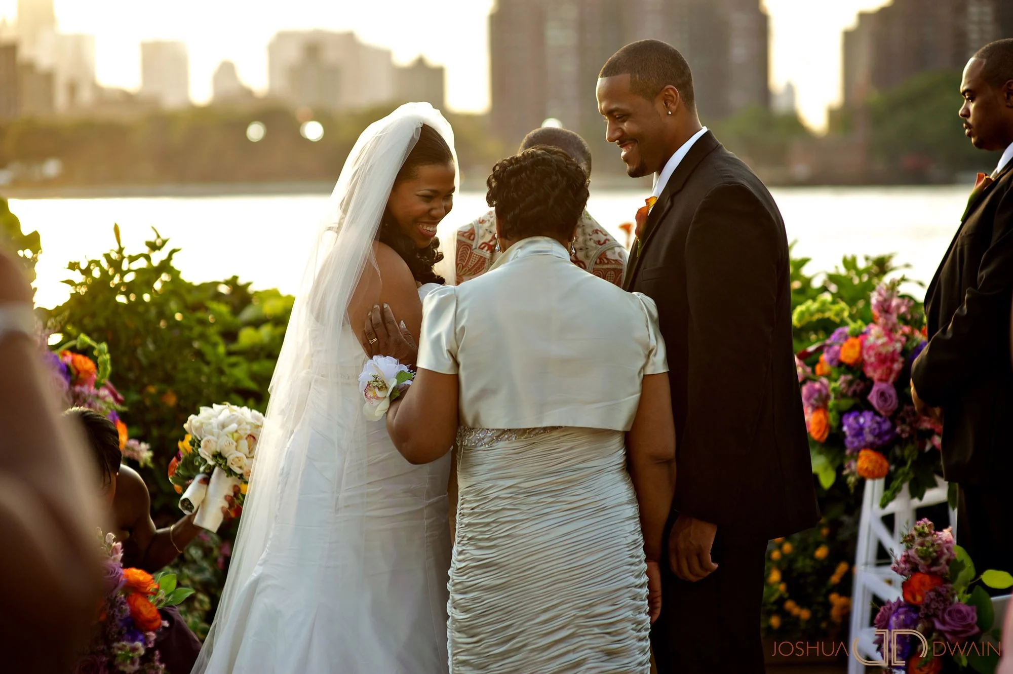 A wedding ceremony taking place outdoors with a bride in a white dress and veil, a groom in a black suit, and family members, surrounded by colorful flowers, against a backdrop of city buildings and water.