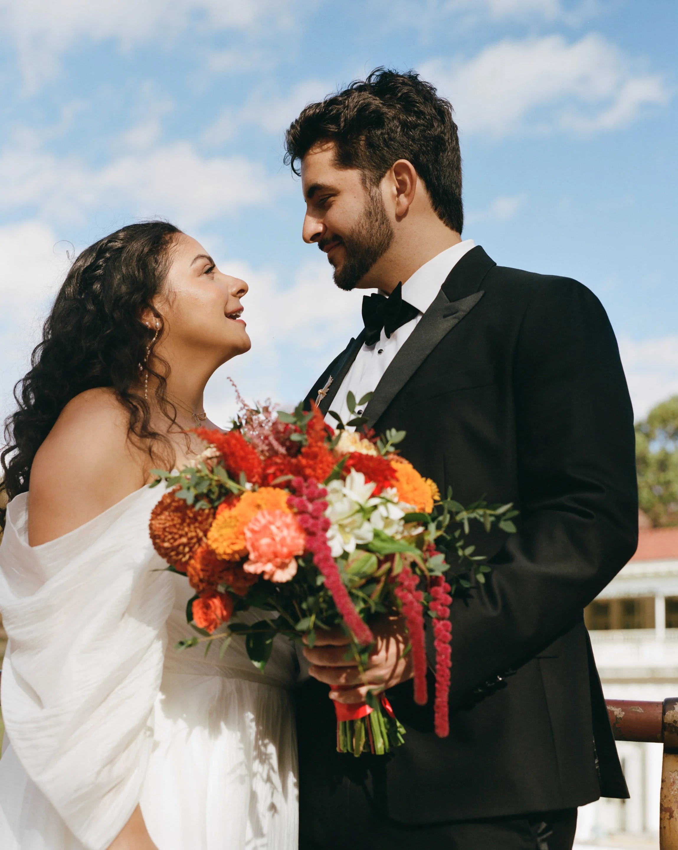 A bride and groom face each other outdoors, with the groom holding a large bouquet of vibrant flowers, during a wedding ceremony under a blue sky with clouds.