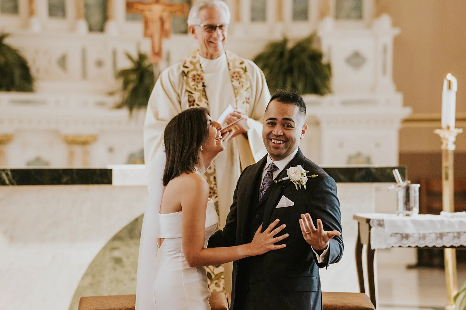 A wedding ceremony inside a church with a bride and groom exchanging vows, a priest officiating, and the bride and groom smiling at each other.