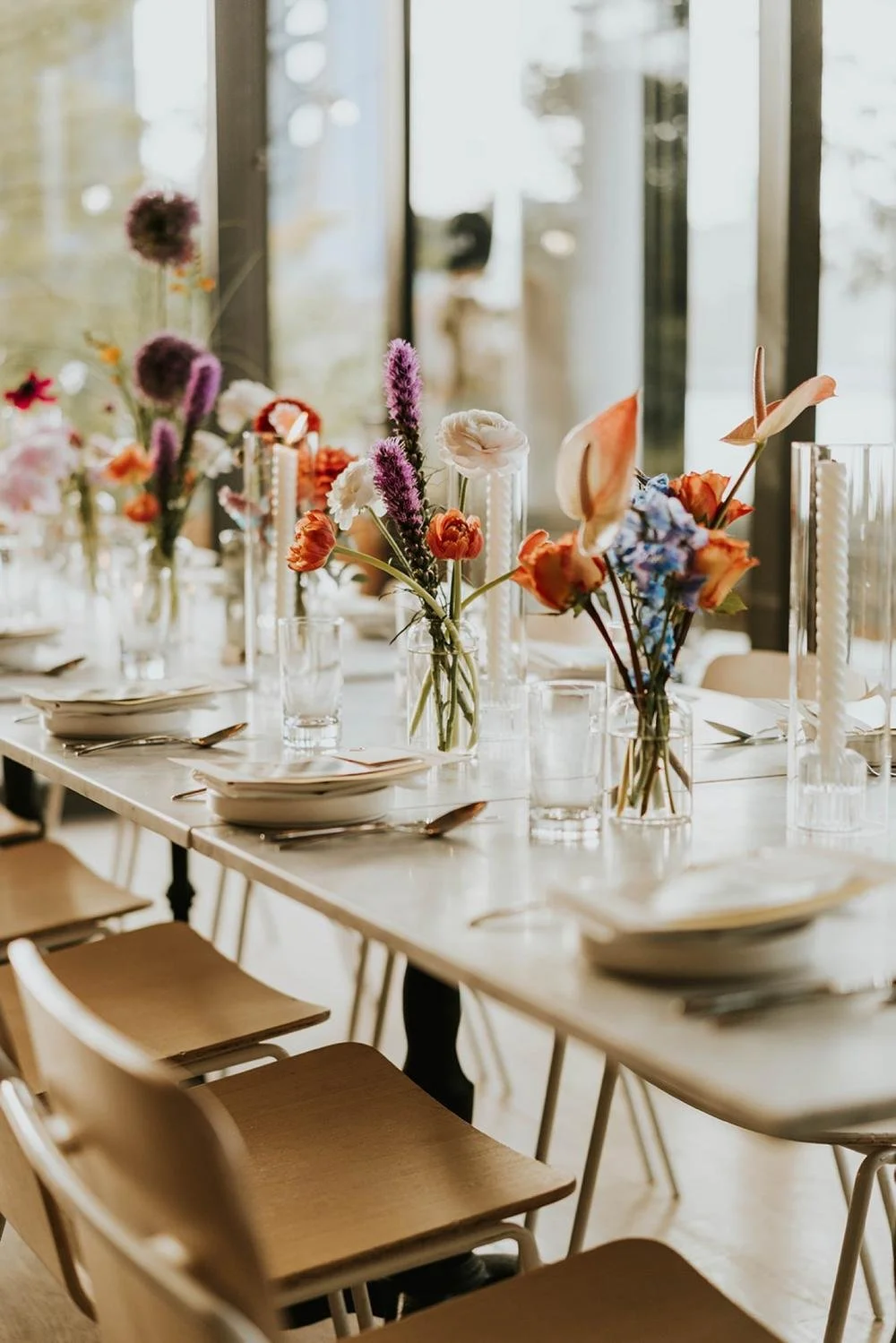 A long dining table decorated with colorful flower arrangements in glass vases, set with plates, silverware, and napkins, in a well-lit room with large windows.