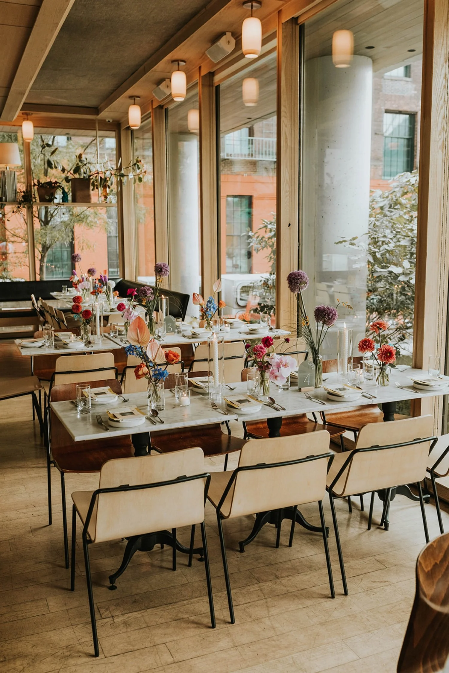 A decorated dining area with long tables set for a celebration or event. The tables are adorned with colorful flower arrangements and candles. Large windows in the background let in natural light, revealing an outdoor urban scene with brick buildings