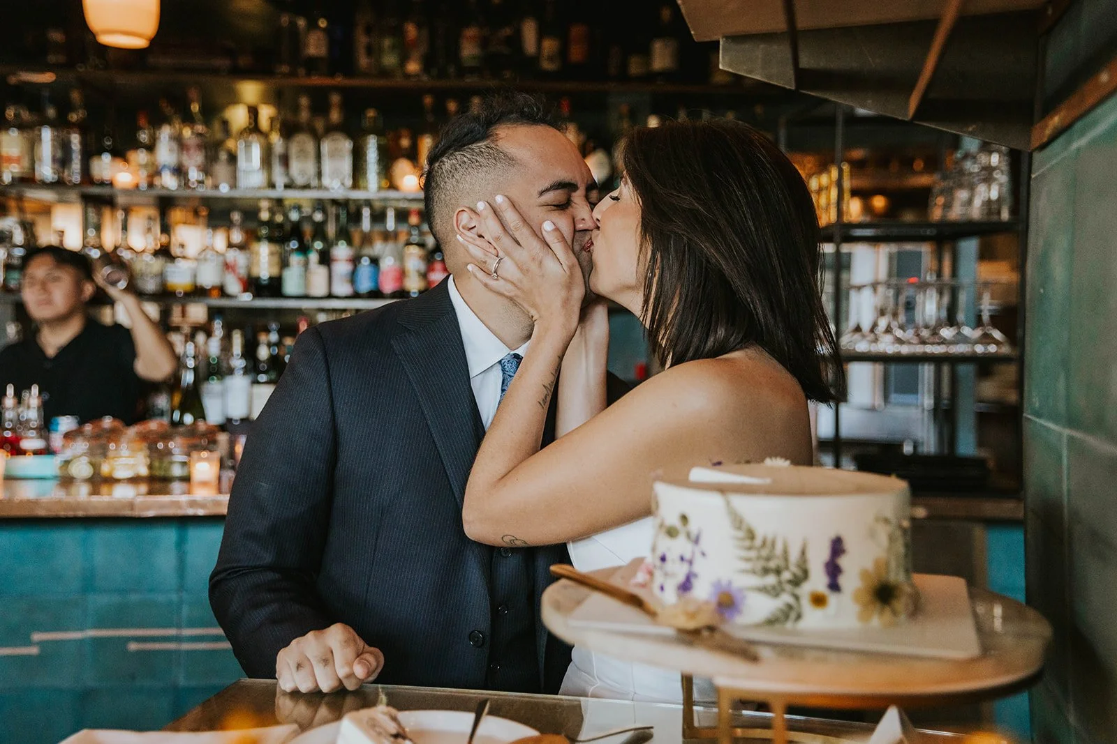 A couple sharing a kiss at a celebration, with a cake in the foreground and a bar with bottles in the background.