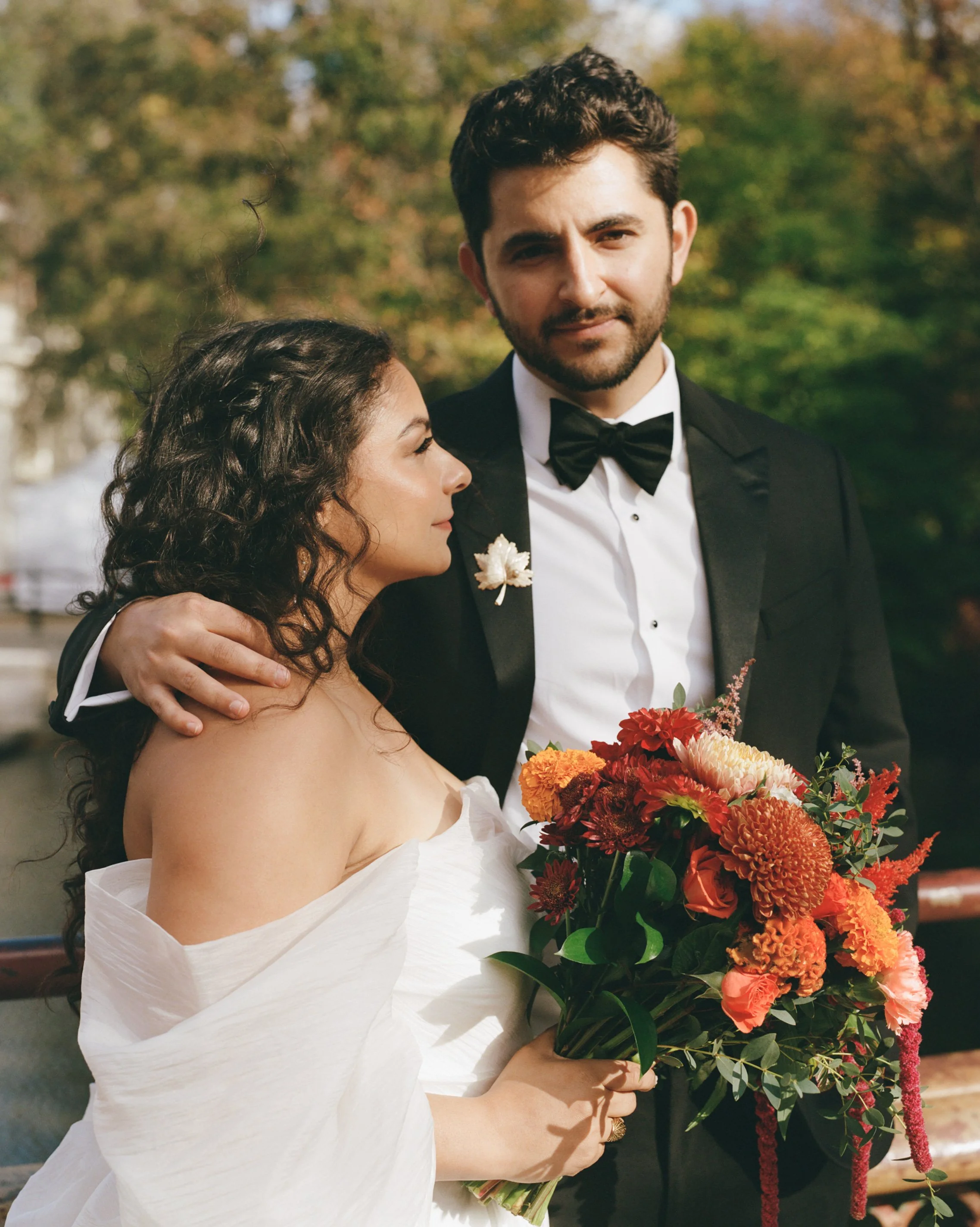 A bride and groom at their wedding, with the bride holding a vibrant bouquet of orange, red, and pink flowers. The groom is dressed in a tuxedo with a bow tie, and they are outdoors with blurred green and autumn-colored trees in the background.