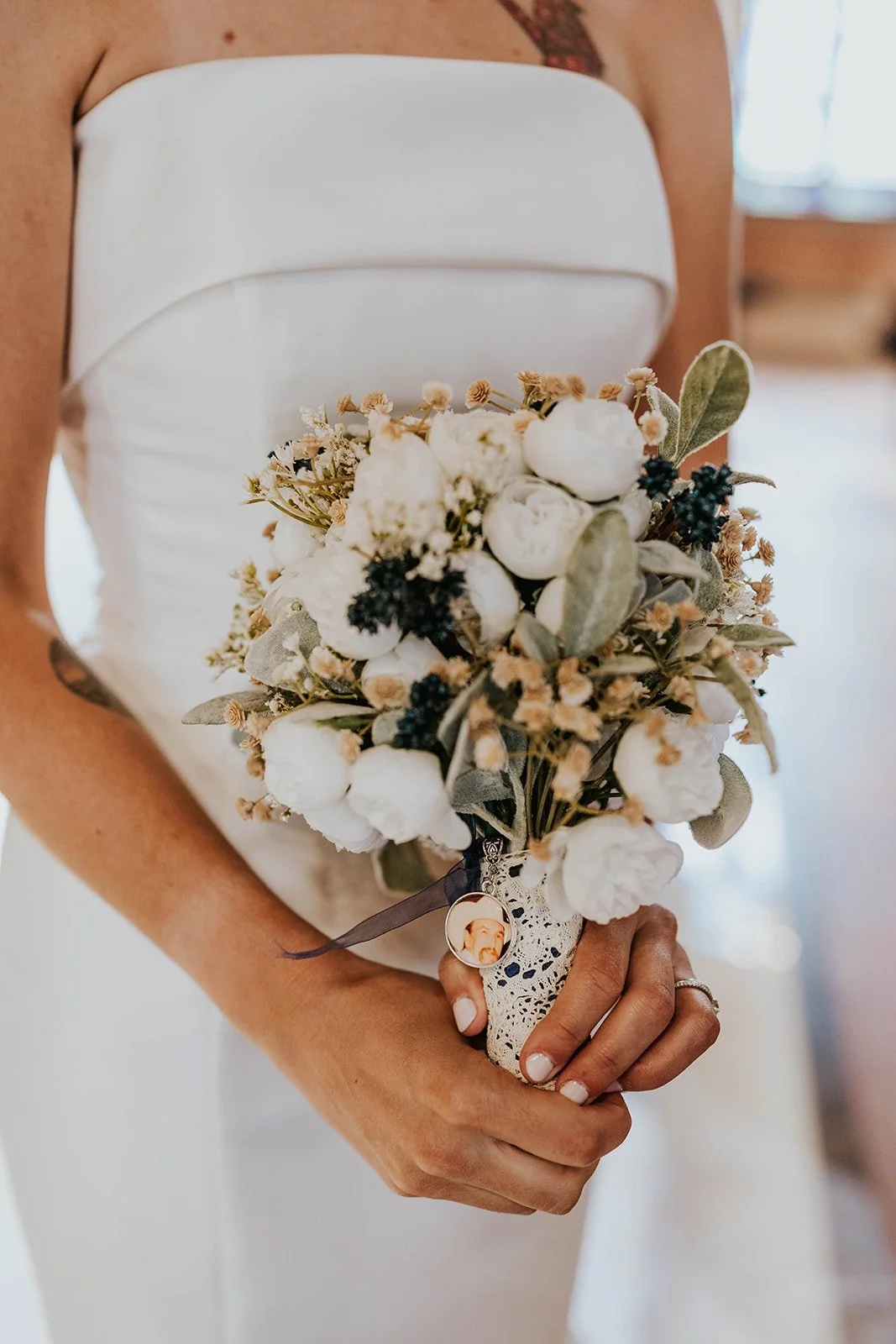 A bride holding a bouquet of white and dark flowers, wrapped with lace and ribbons, with a photo charm attached.
