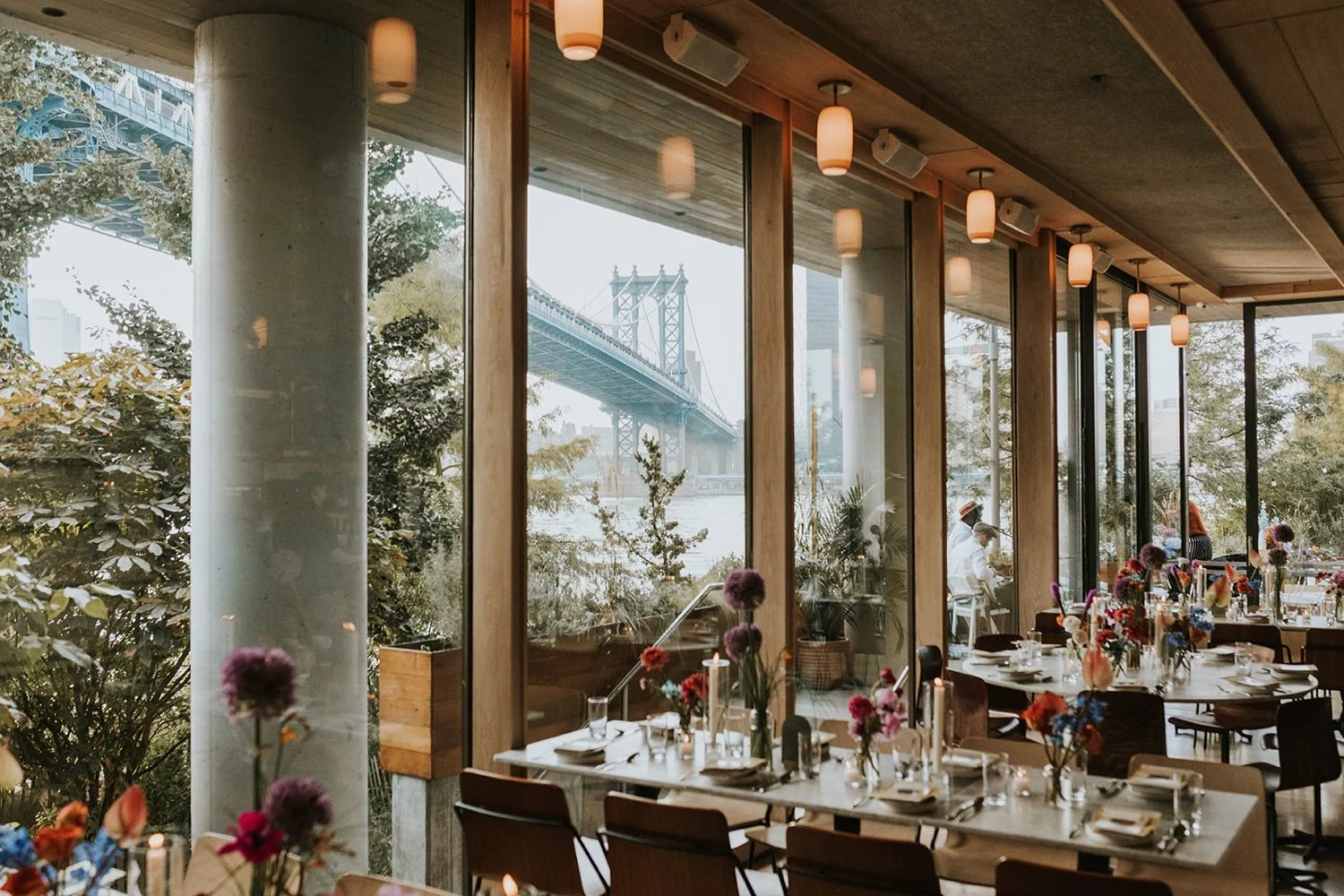 Interior of a restaurant with large glass windows overlooking a bridge and water, decorated with flowers on tables, with some patrons dining.