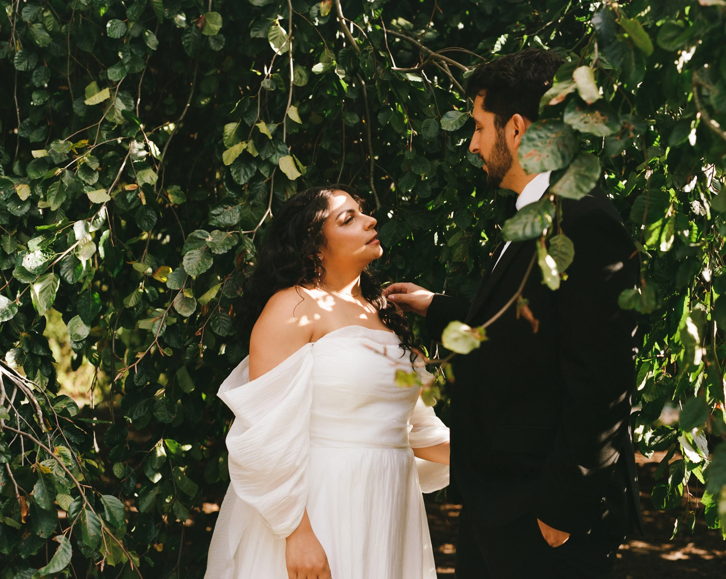 A woman in a white dress and a man in a black suit face each other in front of dense green foliage, possibly during a wedding or special outdoor event.