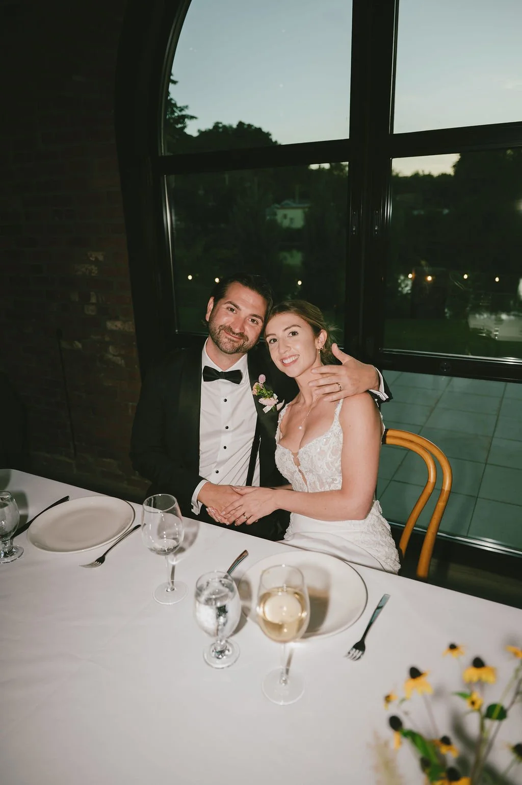 A man in a tuxedo with a bowtie and a woman in a white wedding dress sitting at a dining table, smiling and embracing each other during a wedding celebration indoors, with a large window showing an outdoor landscape in the background.