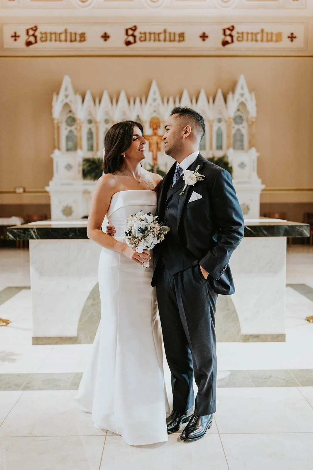 A bride and groom standing close together during their wedding ceremony inside a church, with the bride holding a bouquet of white and dark flowers, and both looking at each other fondly.