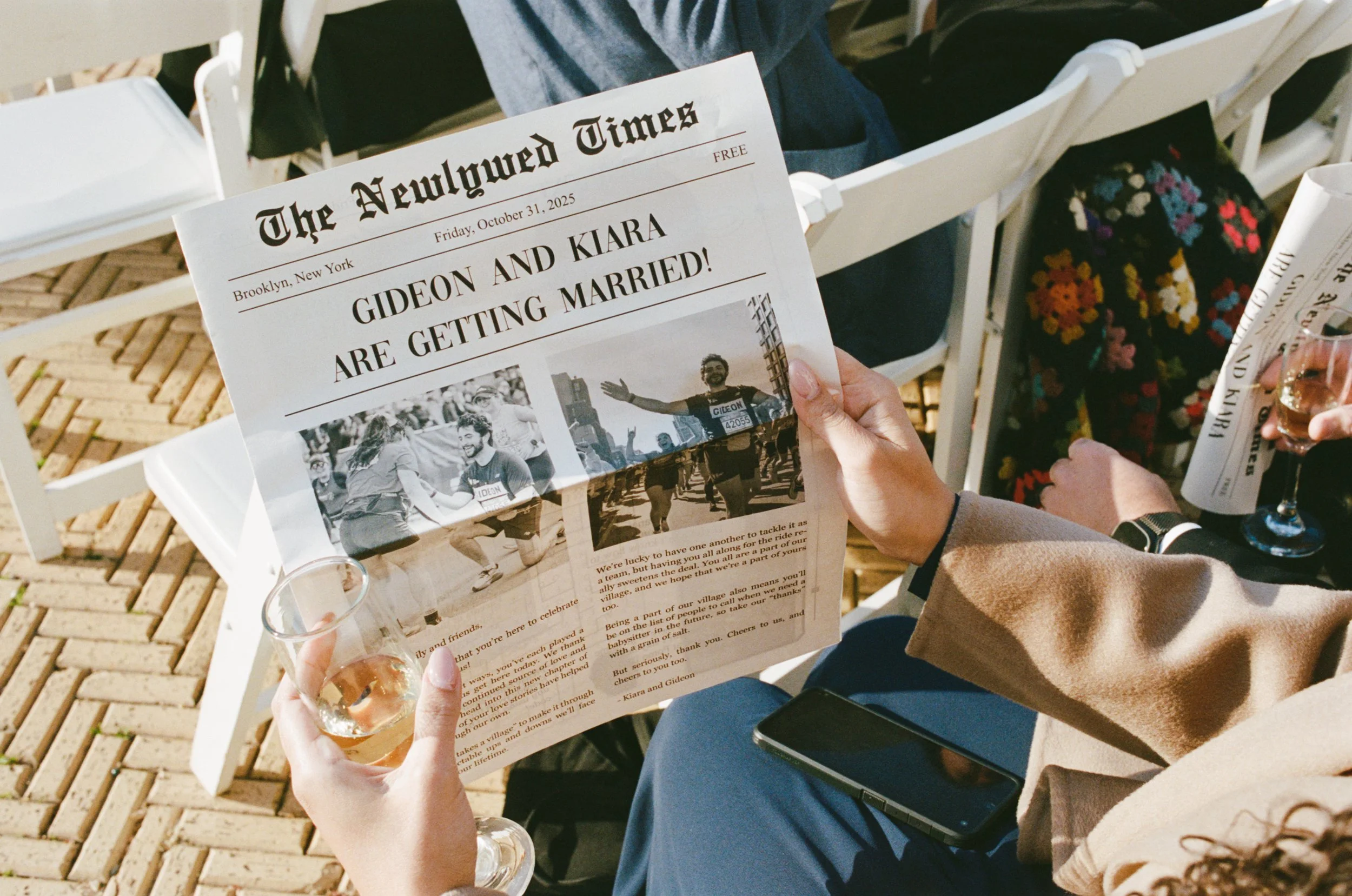A person holding a glass of champagne in one hand and a newspaper with the headline about Gideon and Kiara getting married in the other hand. They are sitting on a white chair outdoors on a brick patio, with some other chairs and people in the backgr