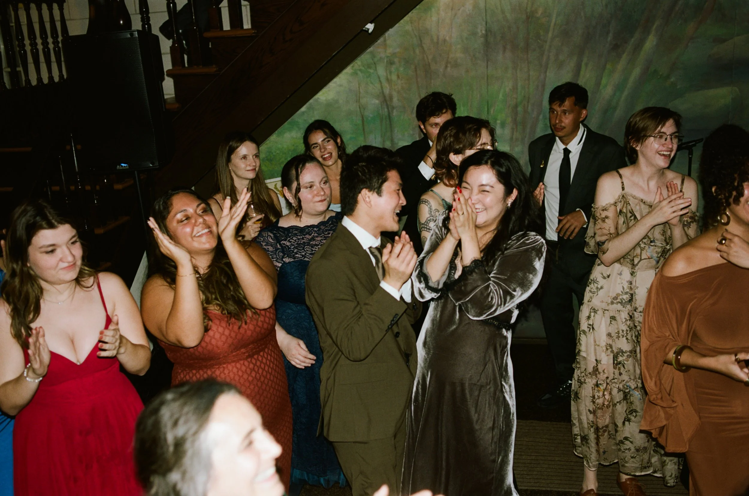 A group of people at a celebration or party, some are smiling and clapping, others are talking and enjoying the moment, with a painted mural background and wooden staircase behind them.