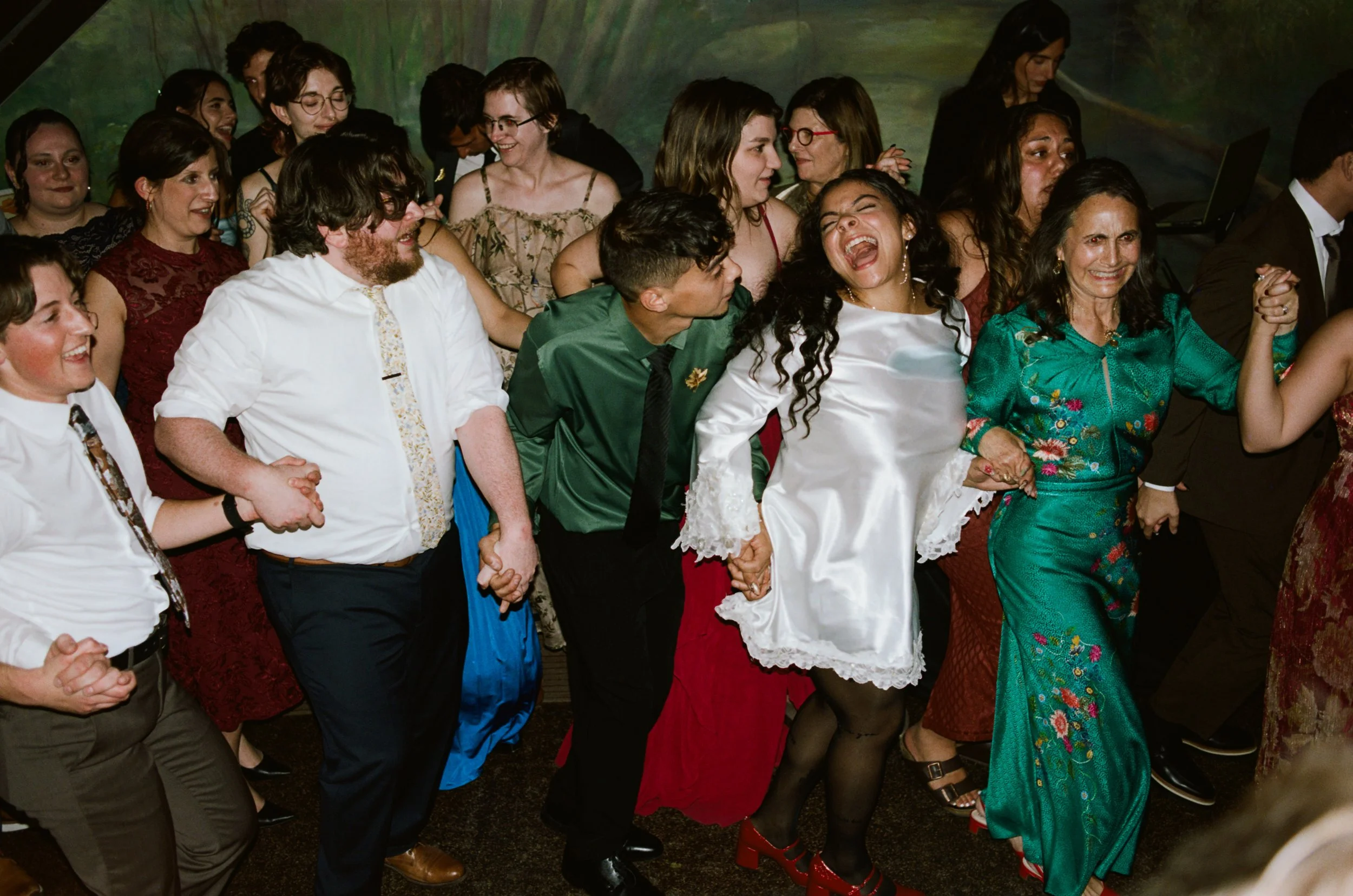A group of people holding hands, laughing, and dancing at a celebration or party, with diverse ages and ethnicities, dressed in semi-formal and traditional clothing.