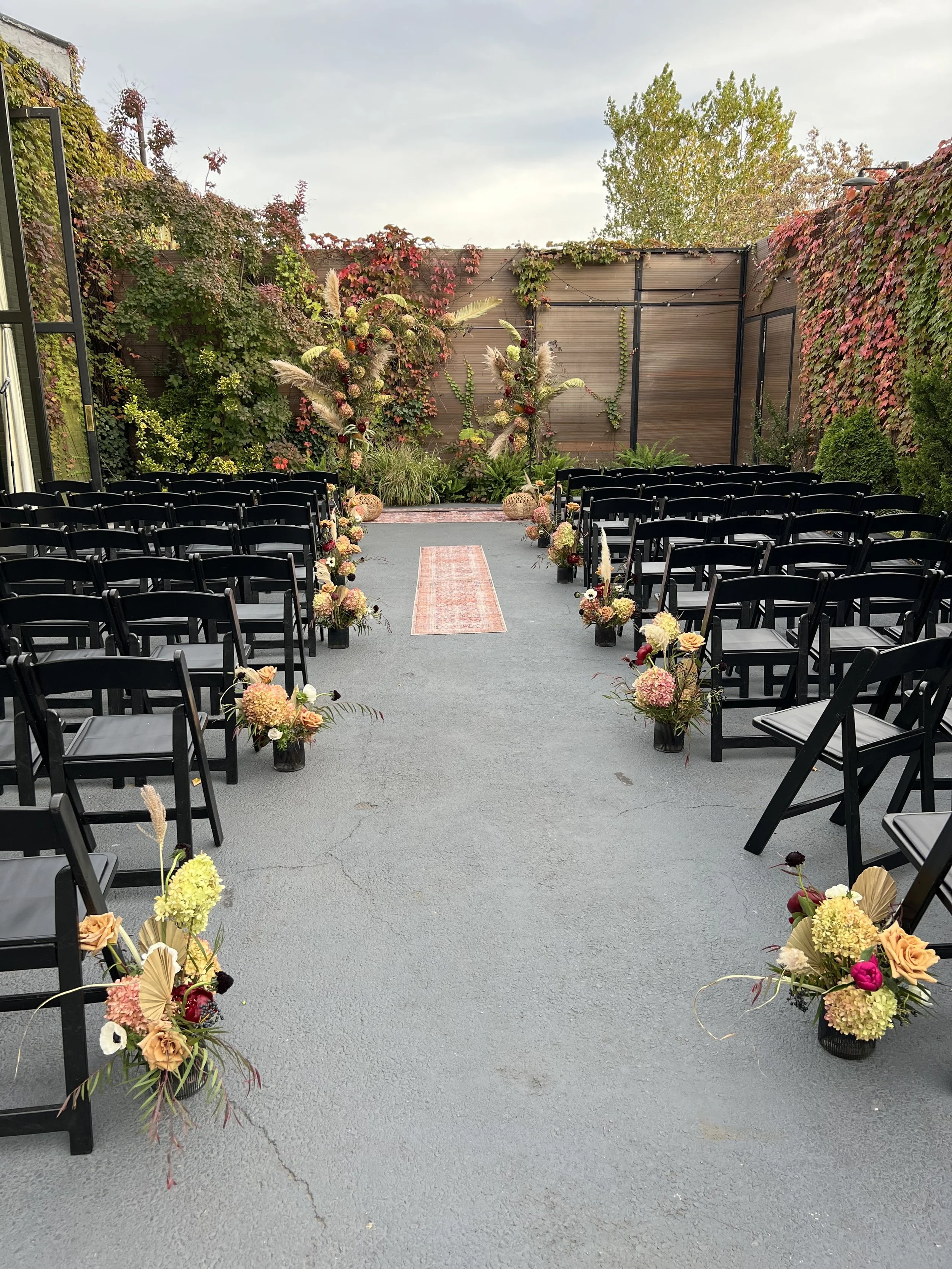 Outdoor wedding ceremony setup with black chairs, floral arrangements, and decorative foliage on a paved area surrounded by garden walls and trees.