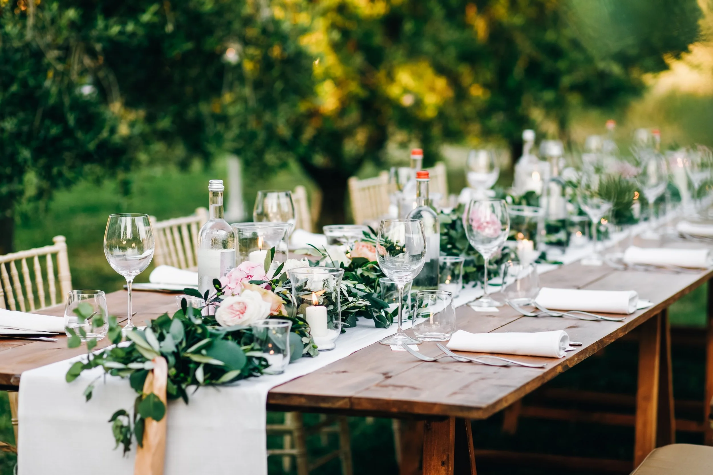 Outdoor dining table decorated with flowers and candles, set for a celebration or event, surrounded by chairs, with trees and greenery in the background.