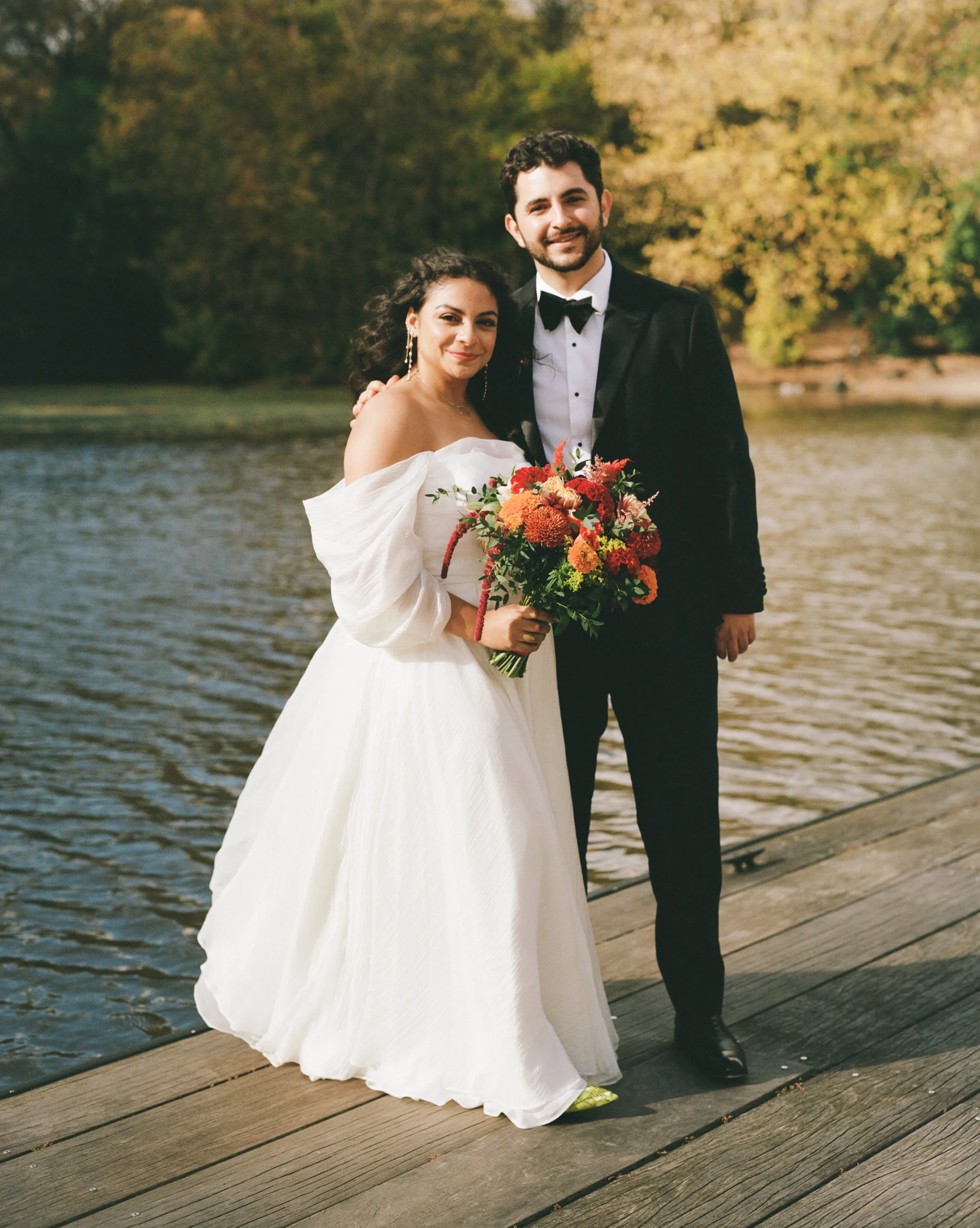 A couple dressed in formal wedding attire standing on a wooden dock by a river, with trees in fall foliage in the background. The woman is holding a large bouquet of colorful flowers, and both are smiling.