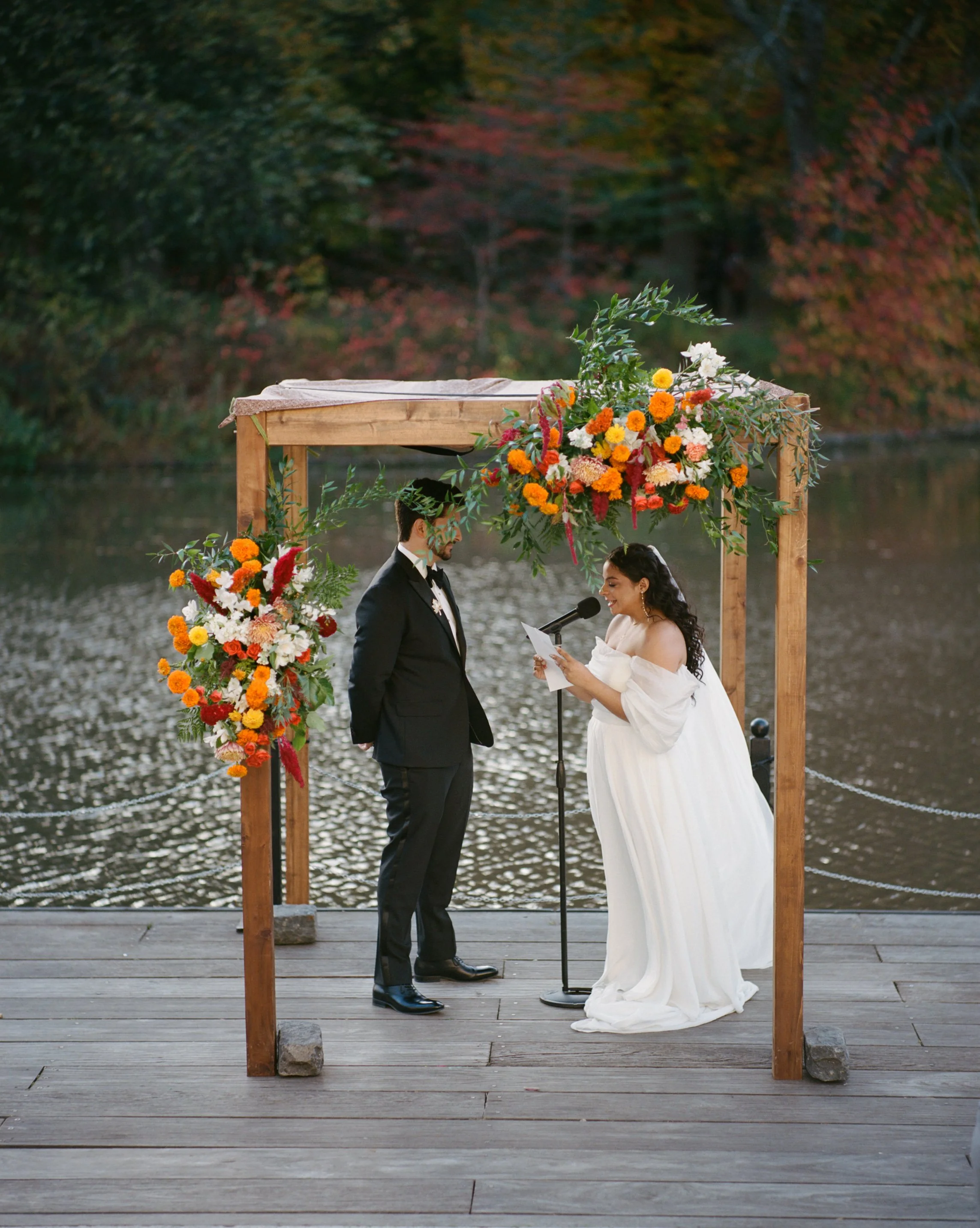 A couple getting married under a decorated arch by a body of water during autumn, with trees showing fall colors in the background.