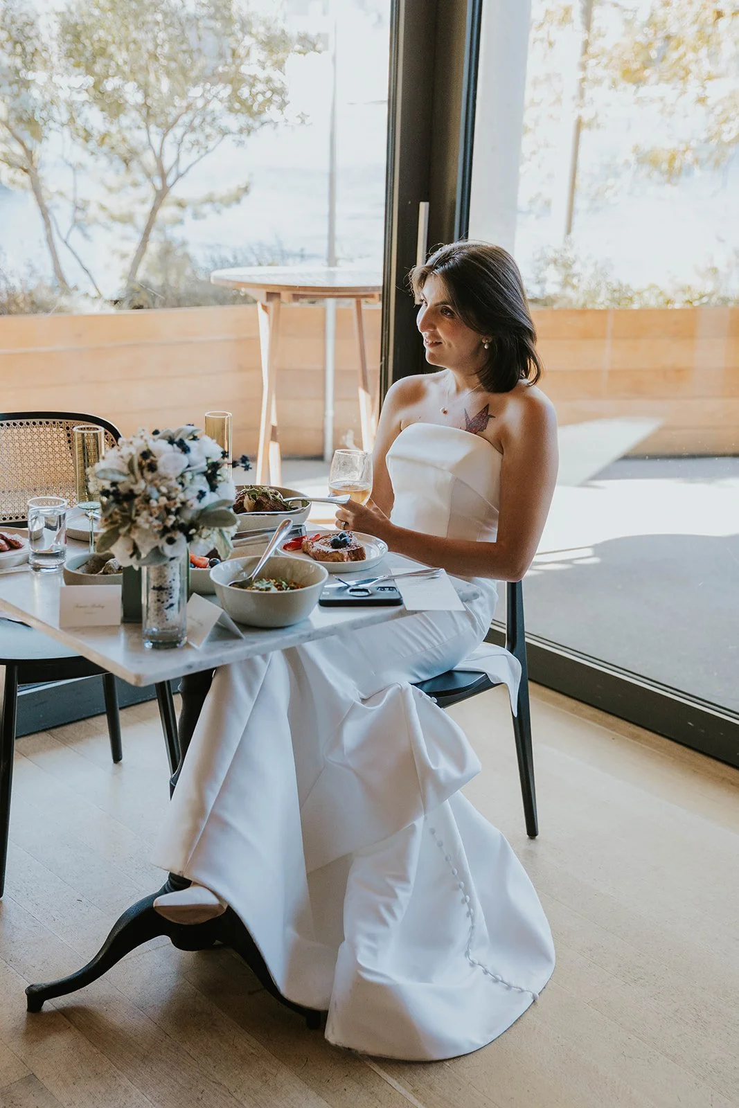 A woman in a white strapless dress sitting at a table set for a meal, holding a glass of rosé, with a flower arrangement and food on the table, near a large glass door with sunlight streaming in.