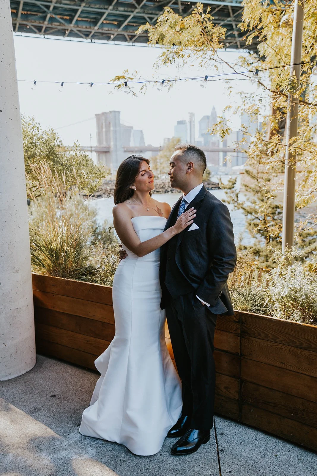 A bride in a white wedding dress and a groom in a black suit are standing close together outdoors with the Brooklyn Bridge and New York City skyline in the background.