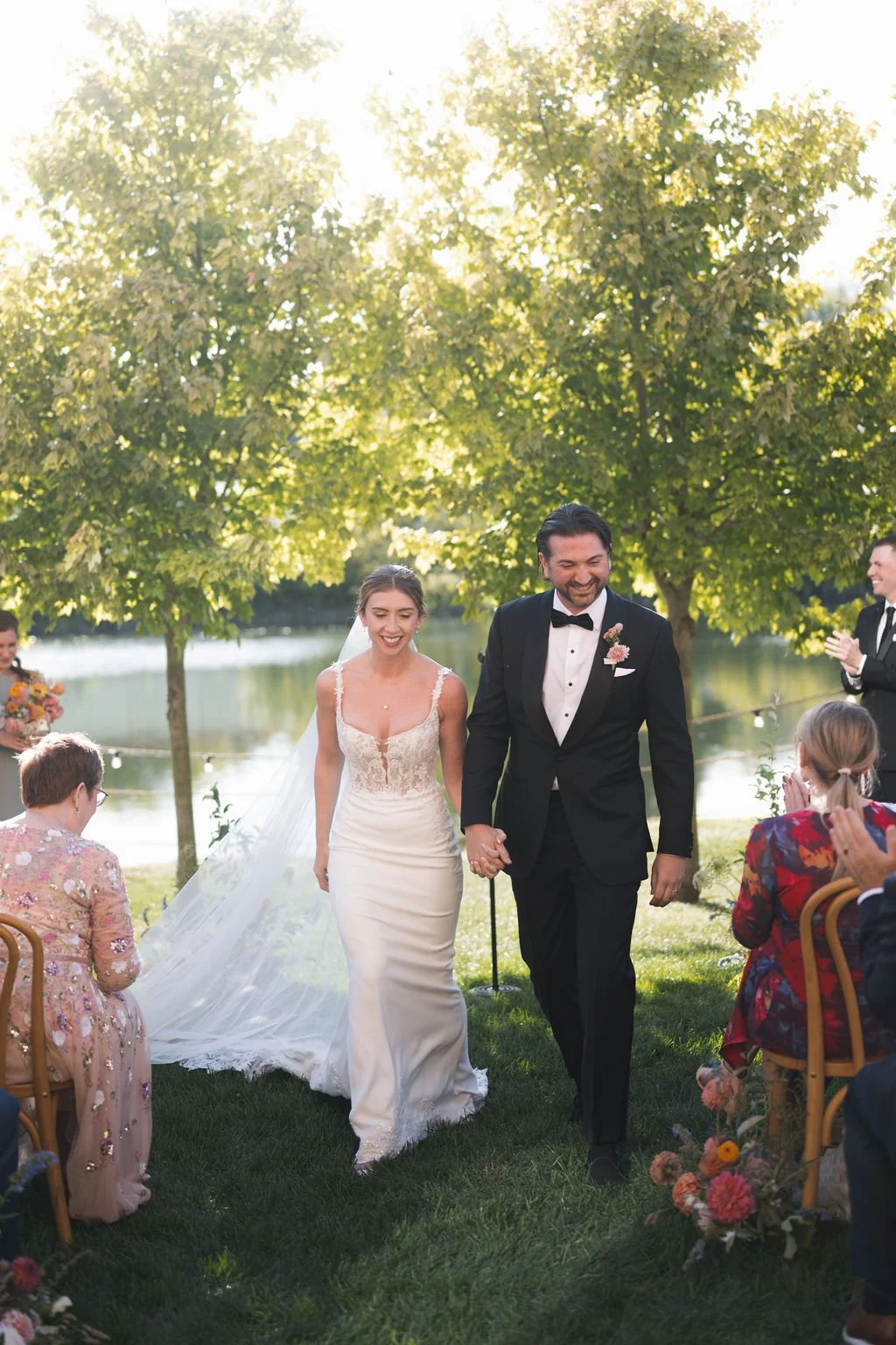 A bride and groom walking hand in hand outdoors during their wedding ceremony under green trees, with guests seated and clapping, on a sunny day by a body of water.