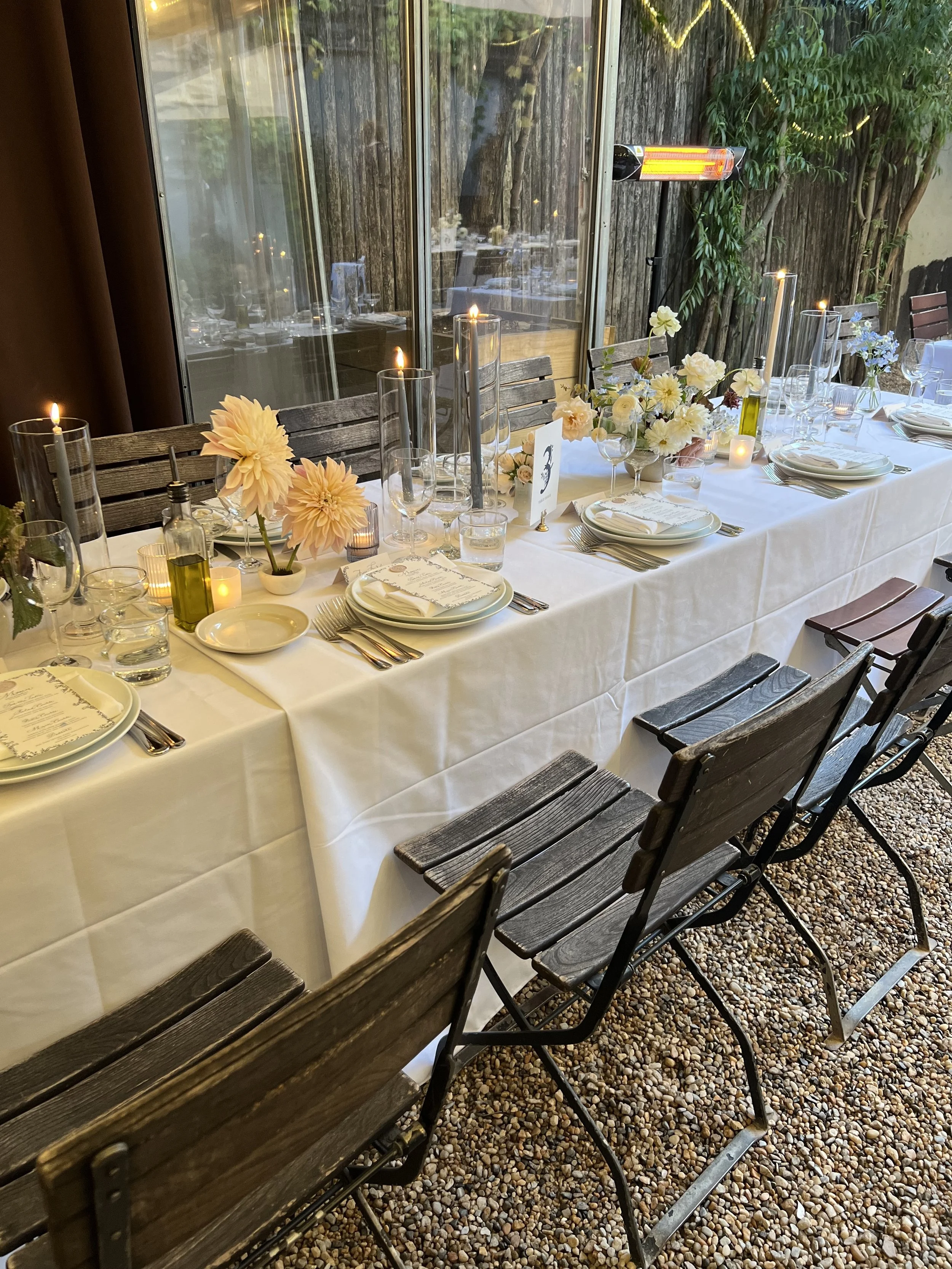 A long rectangular dining table set for a meal with floral centerpieces, candles, and dinnerware; outdoors under a glass roof with natural light and greenery.