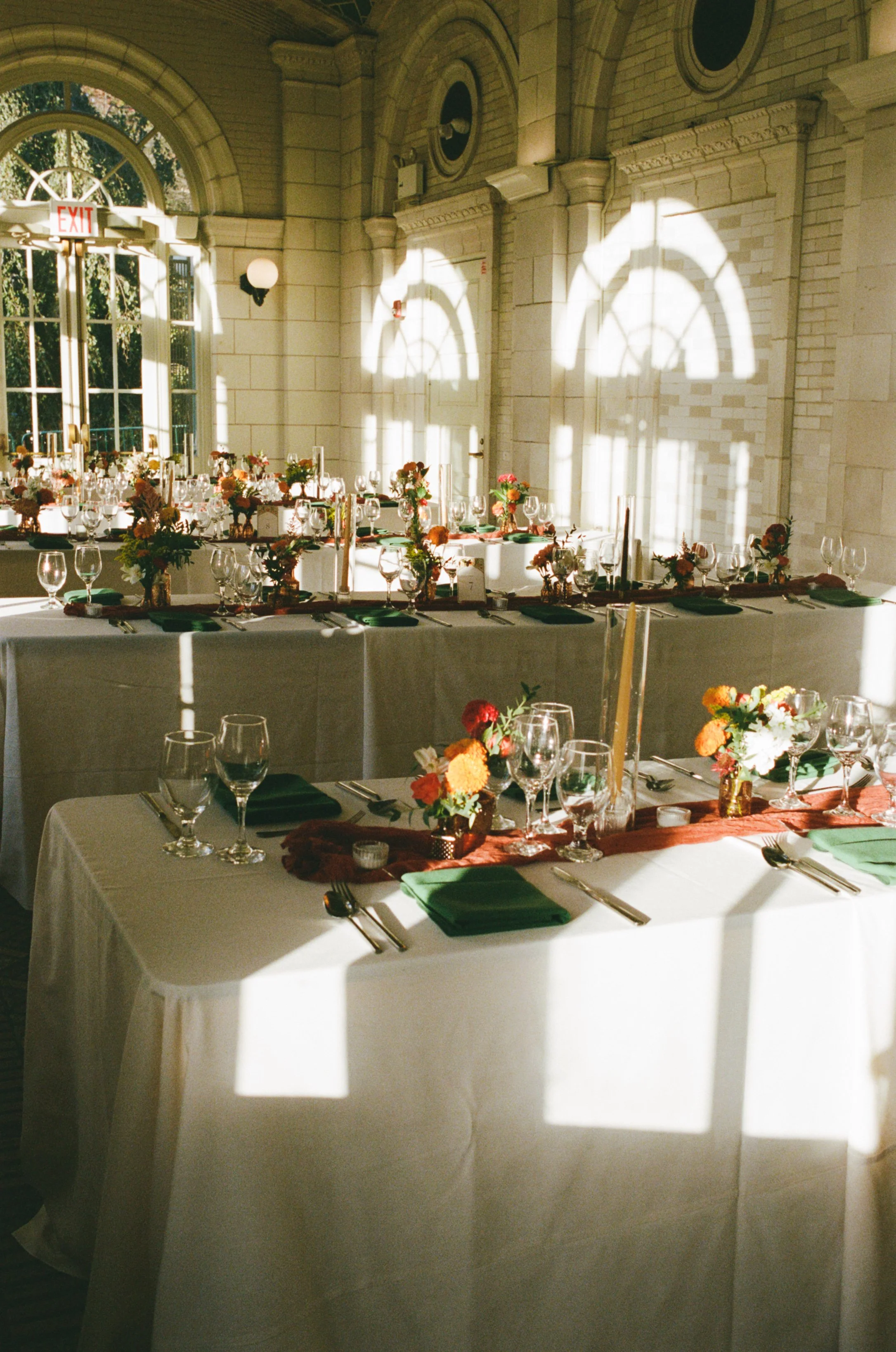 Elegant banquet table setup with floral centerpieces, green napkins, and glassware inside a sunlit brick-walled hall