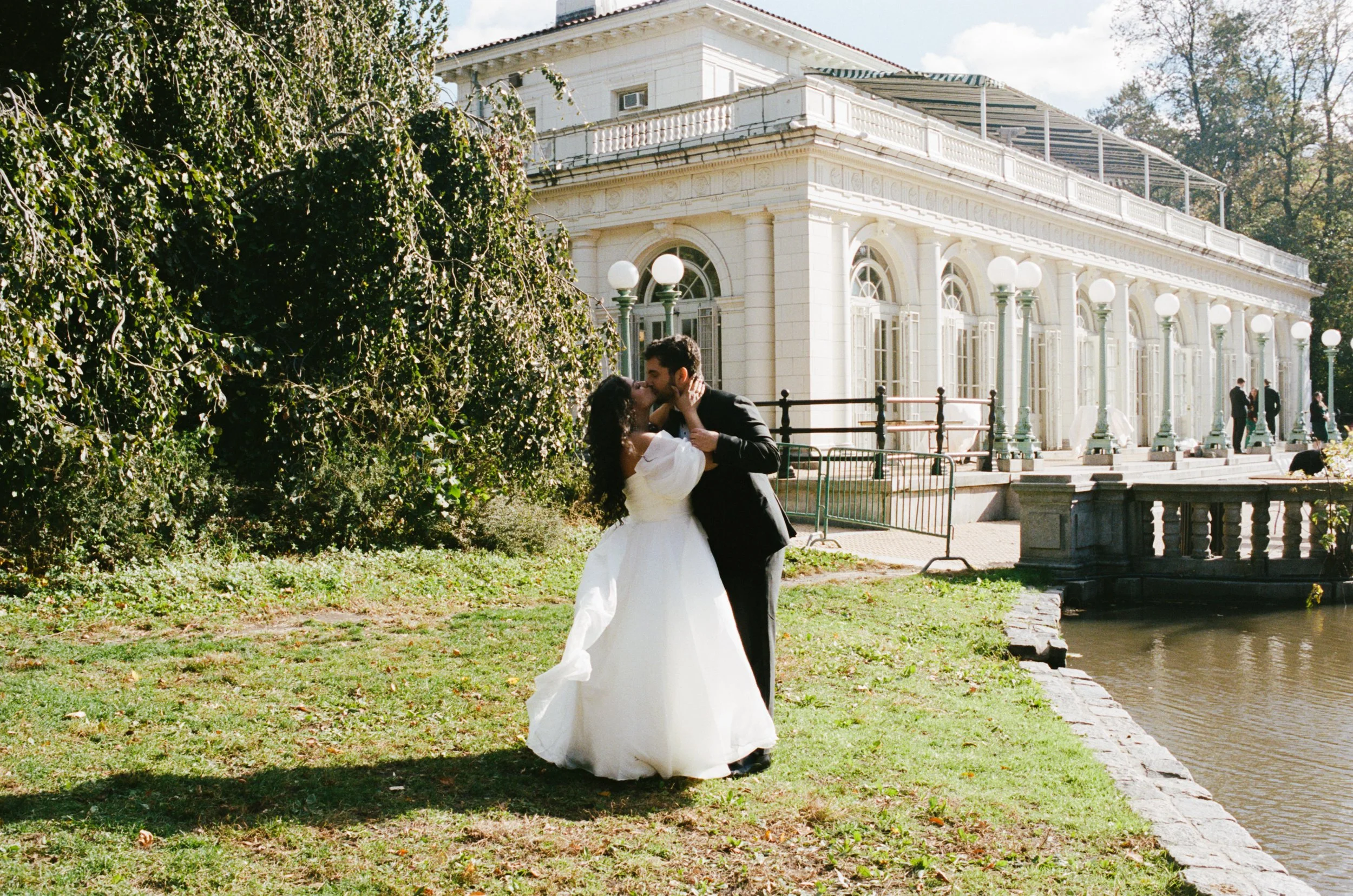A newly married couple kissing outdoors beside a historic white building with large windows and a porch, surrounded by trees and greenery on a sunny day.