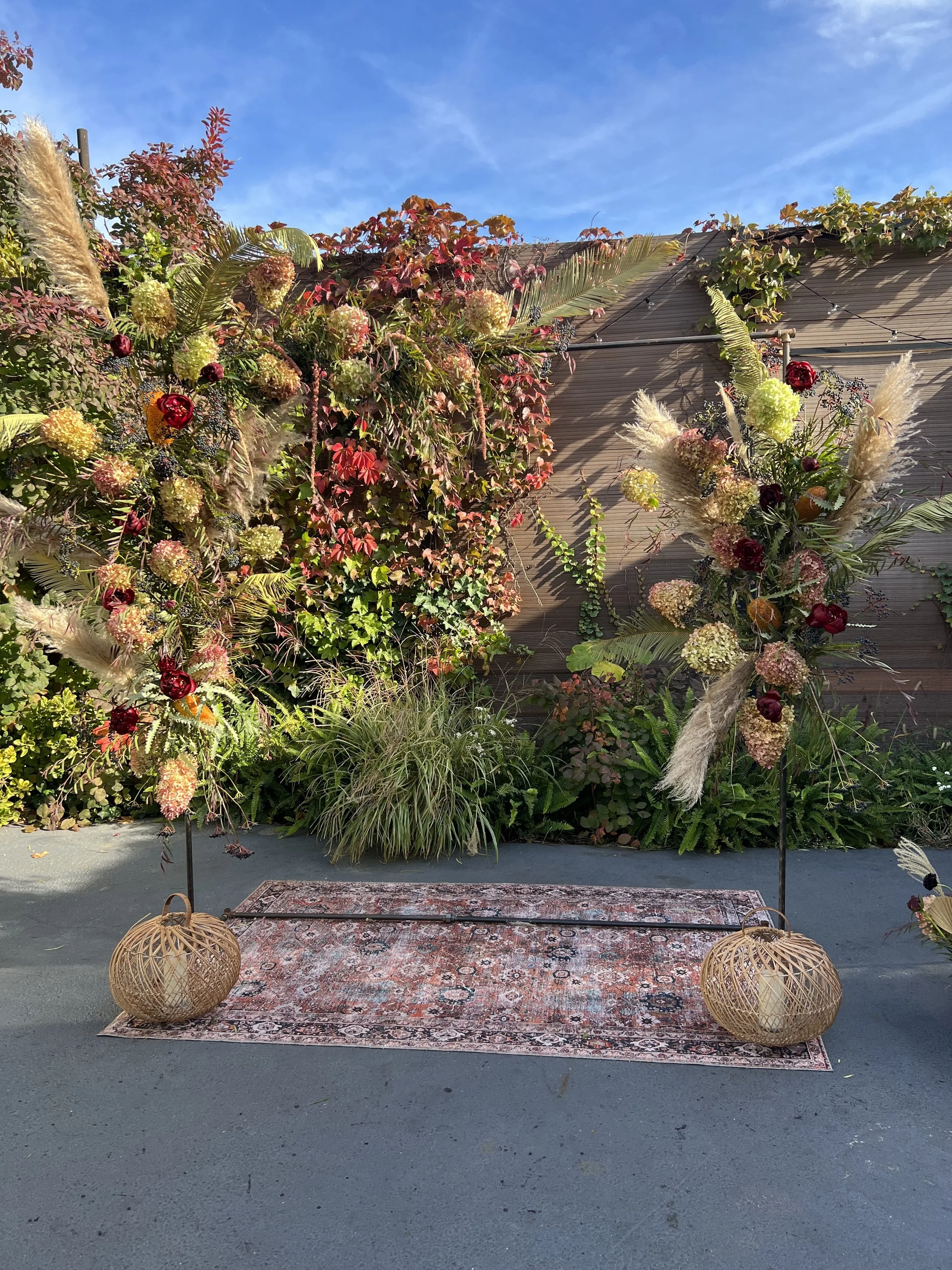Outdoor scene with a floral display set against a wooden wall, decorated with hanging plants, lanterns, and a patterned rug on the ground.