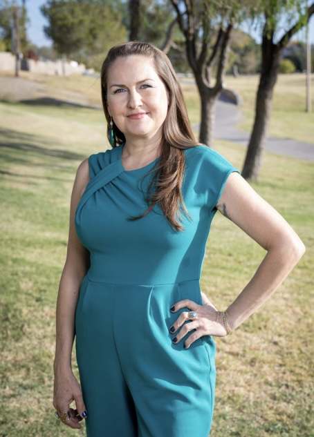 A woman with long brown hair wearing a teal dress standing outdoors in a park with trees and grass, smiling at the camera.
