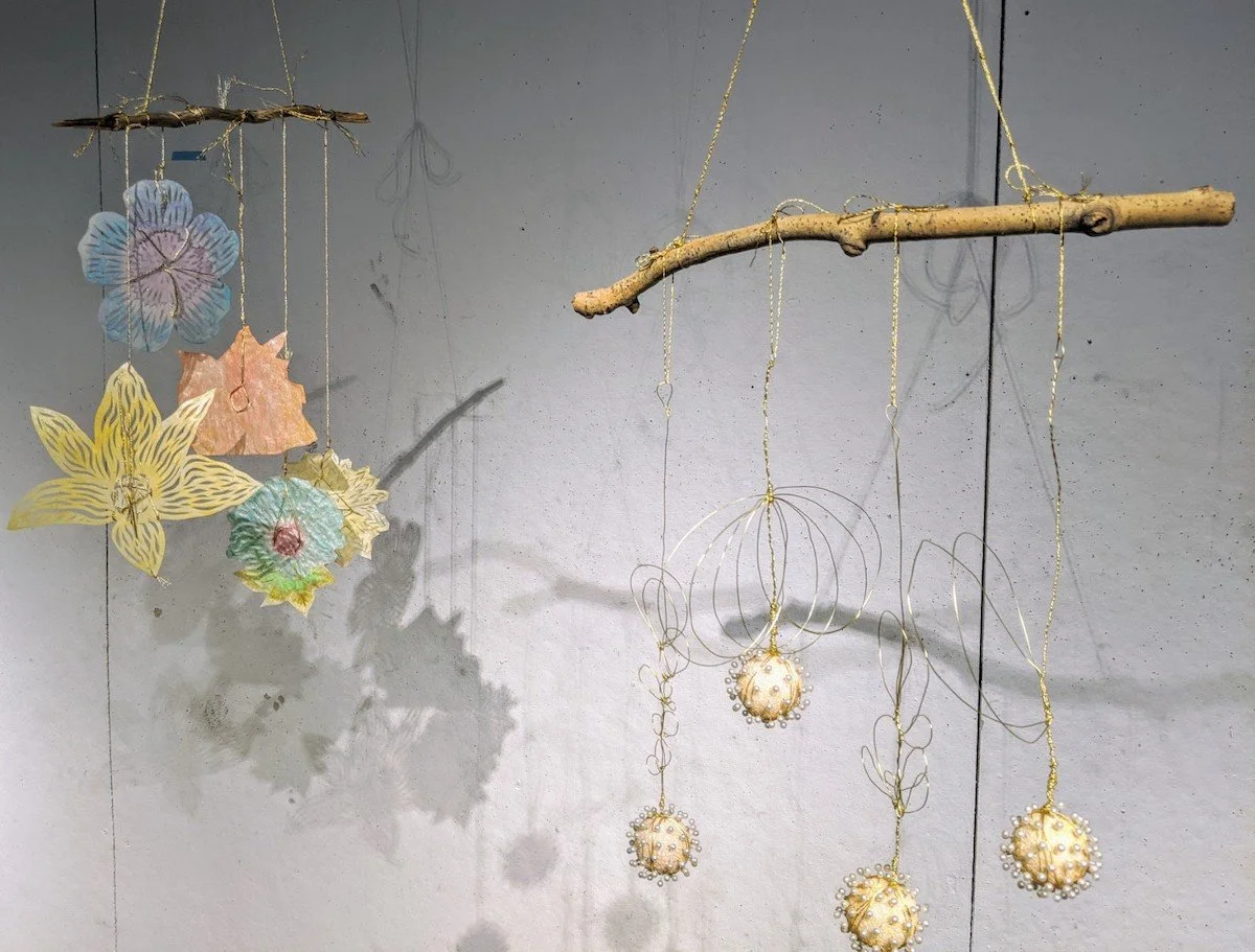 Decorative hanging mobile with paper flowers and cutouts on clouds and a star, suspended from tree branches with gold string, shadow cast on gray wall.