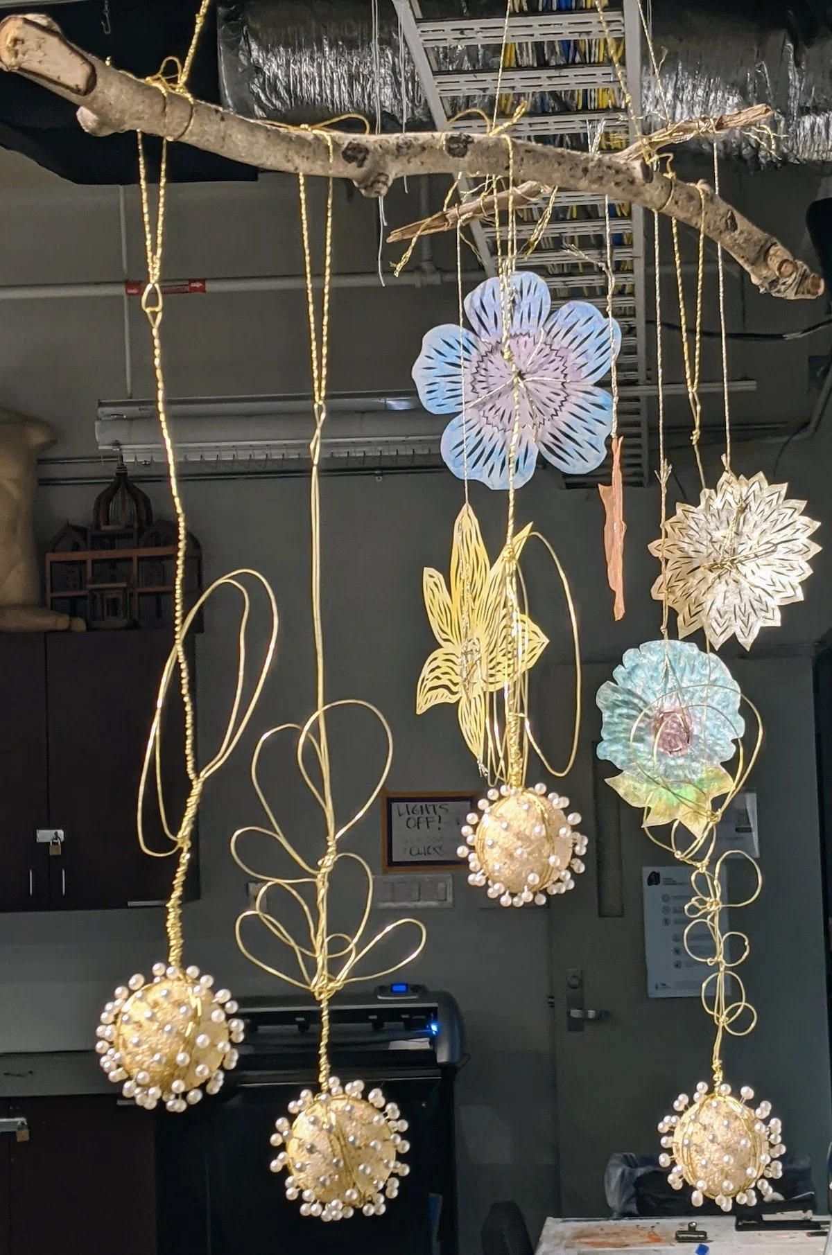 Decorative hanging ornaments made from wire, paper, and lights, suspended from a tree branch in an indoor space.