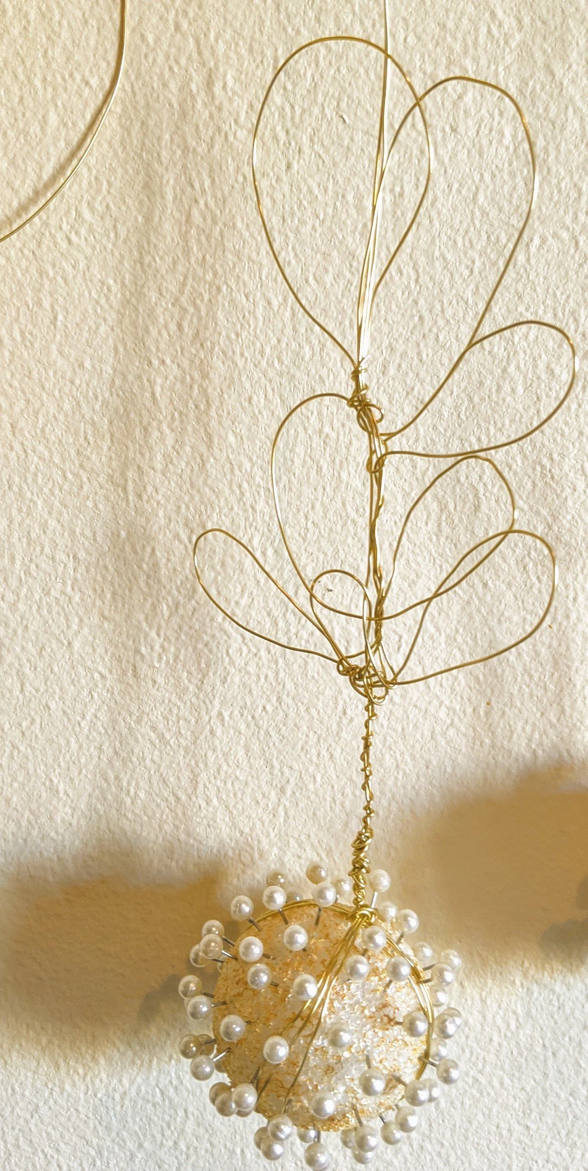 Decorative gold wire sculpture with pearl accents shaped like a heart and abstract forms, hanging against a textured wall.