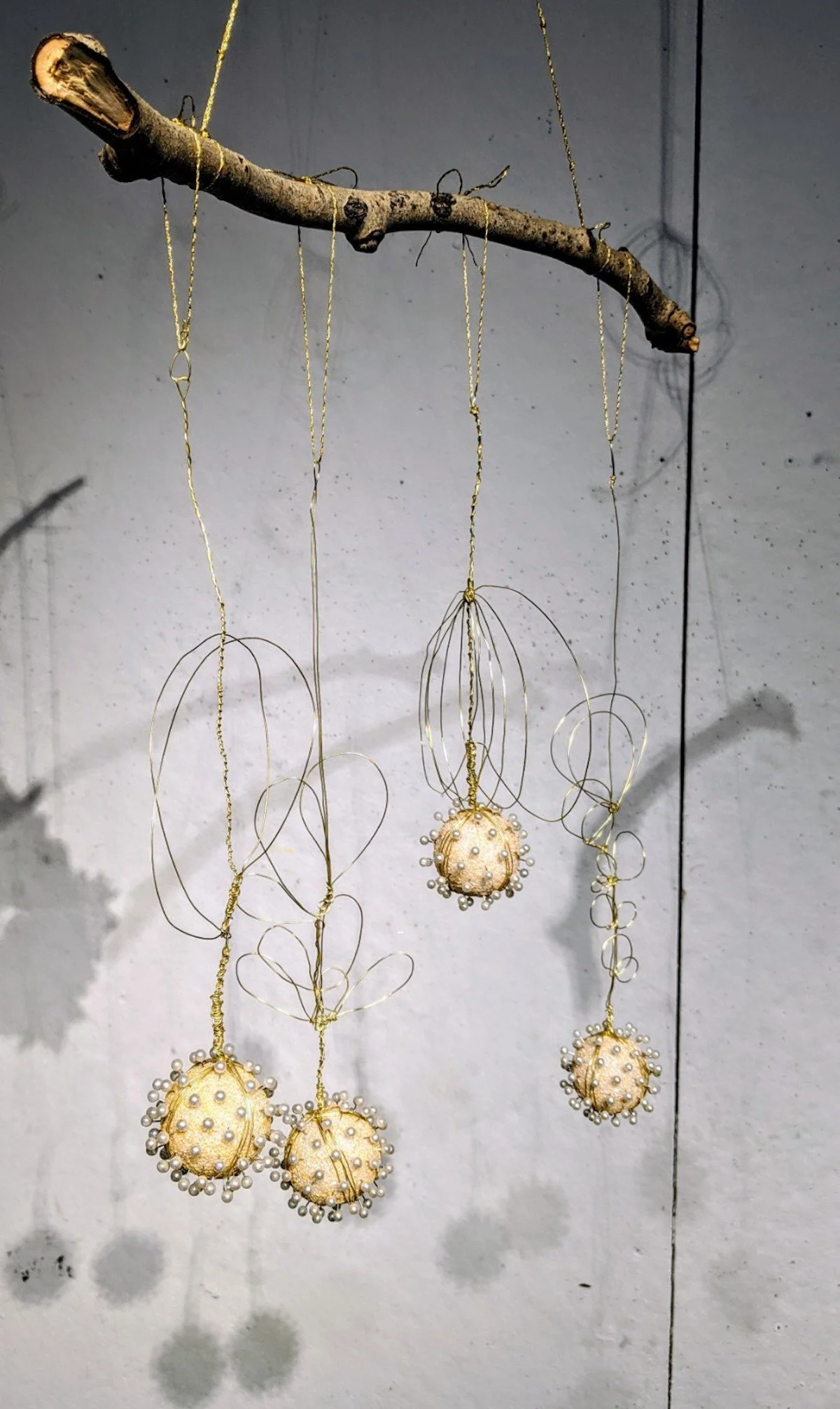 Decorative ornaments hanging from a branch, made with pearl beads and wire, suspended against a plain wall.