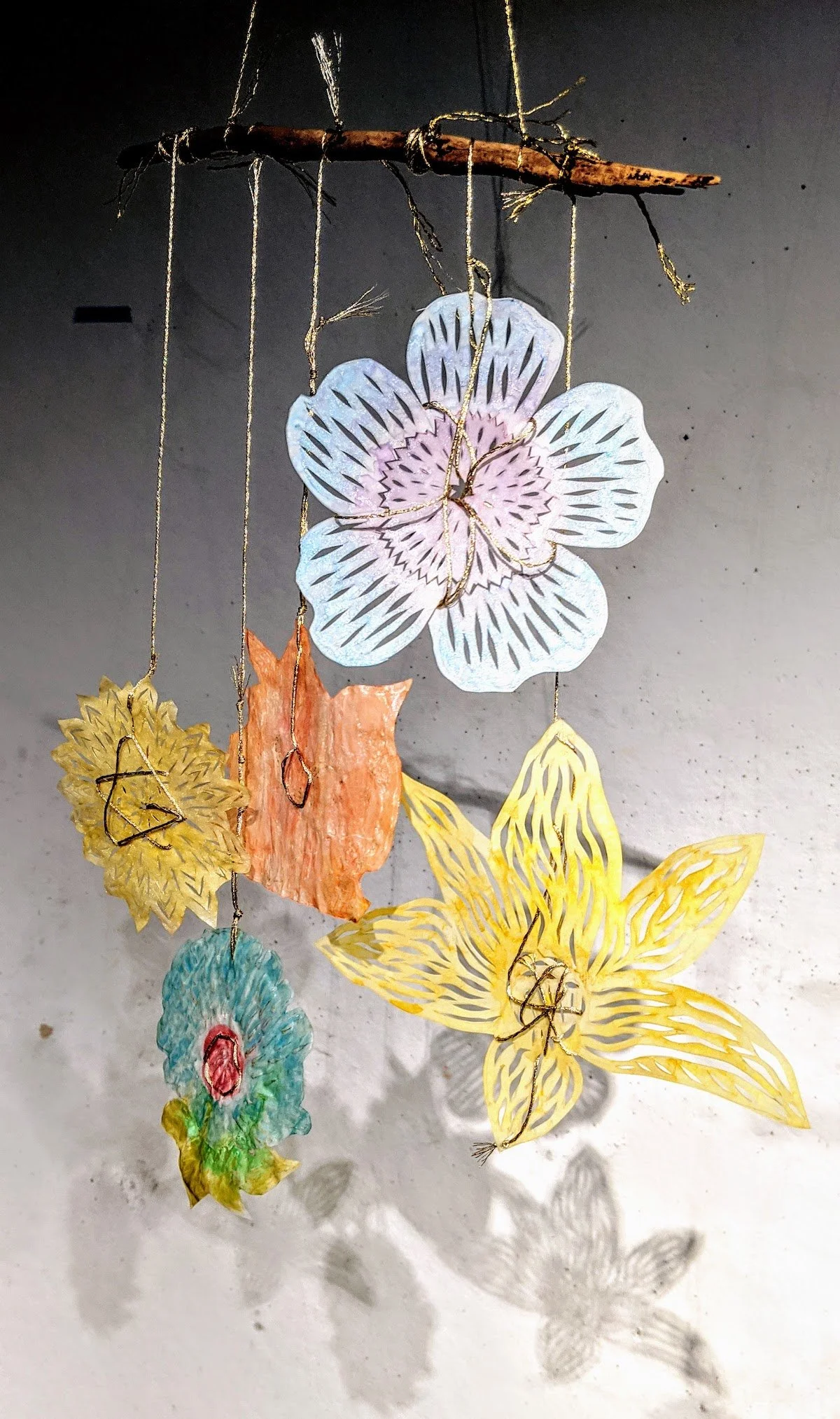 Colorful paper flower mobile hanging from a stick with strings, casting detailed shadow on wall.