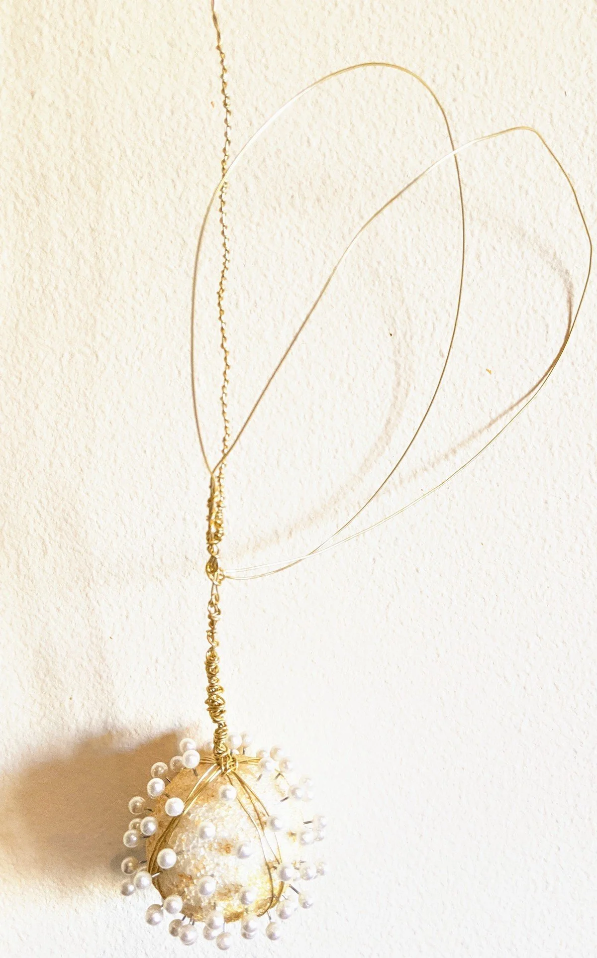 Decorative ornament with white beads and wire, hanging on a white textured wall, with wire loops above it.
