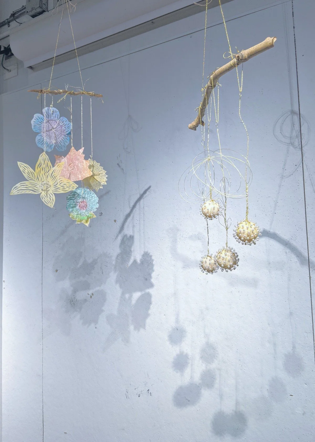 Decorative hanging ornaments made from paper, wood, and beads, featuring flowers and spherical shapes, cast shadows on a plain wall.