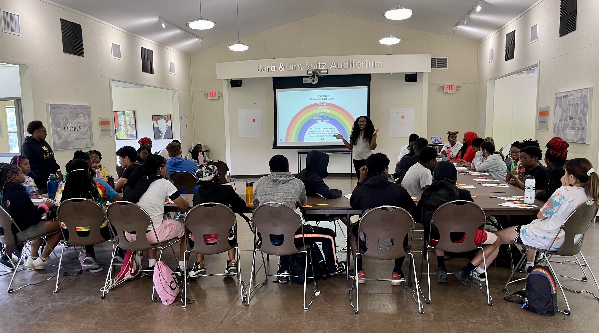 A classroom or workshop setting with a diverse group of students seated at tables, attentively listening to a female presenter at the front of the room. A large screen behind her displays a colorful rainbow infographic and text, with the room labeled 'Barb & Jim Saitz Auditorium'.