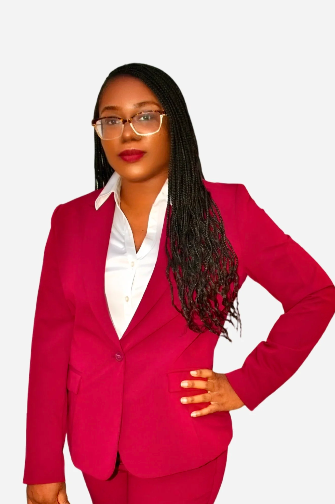 A woman with long, braided black hair, wearing glasses, a white shirt, and a pink suit, standing with her hand on her hip against a plain background.
