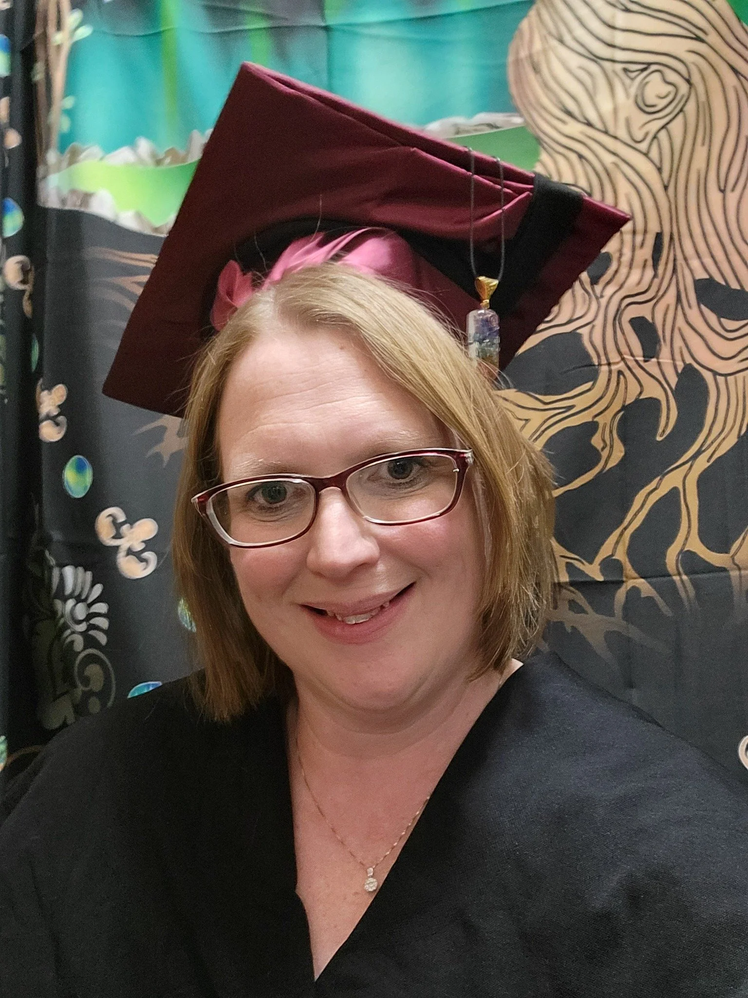 A smiling woman with glasses and shoulder-length brown hair wearing a black top and a necklace, posing in front of a colorful background with a large illustration of an animal with a distinctive patterned face and a person in a pink graduation cap and gown behind her.